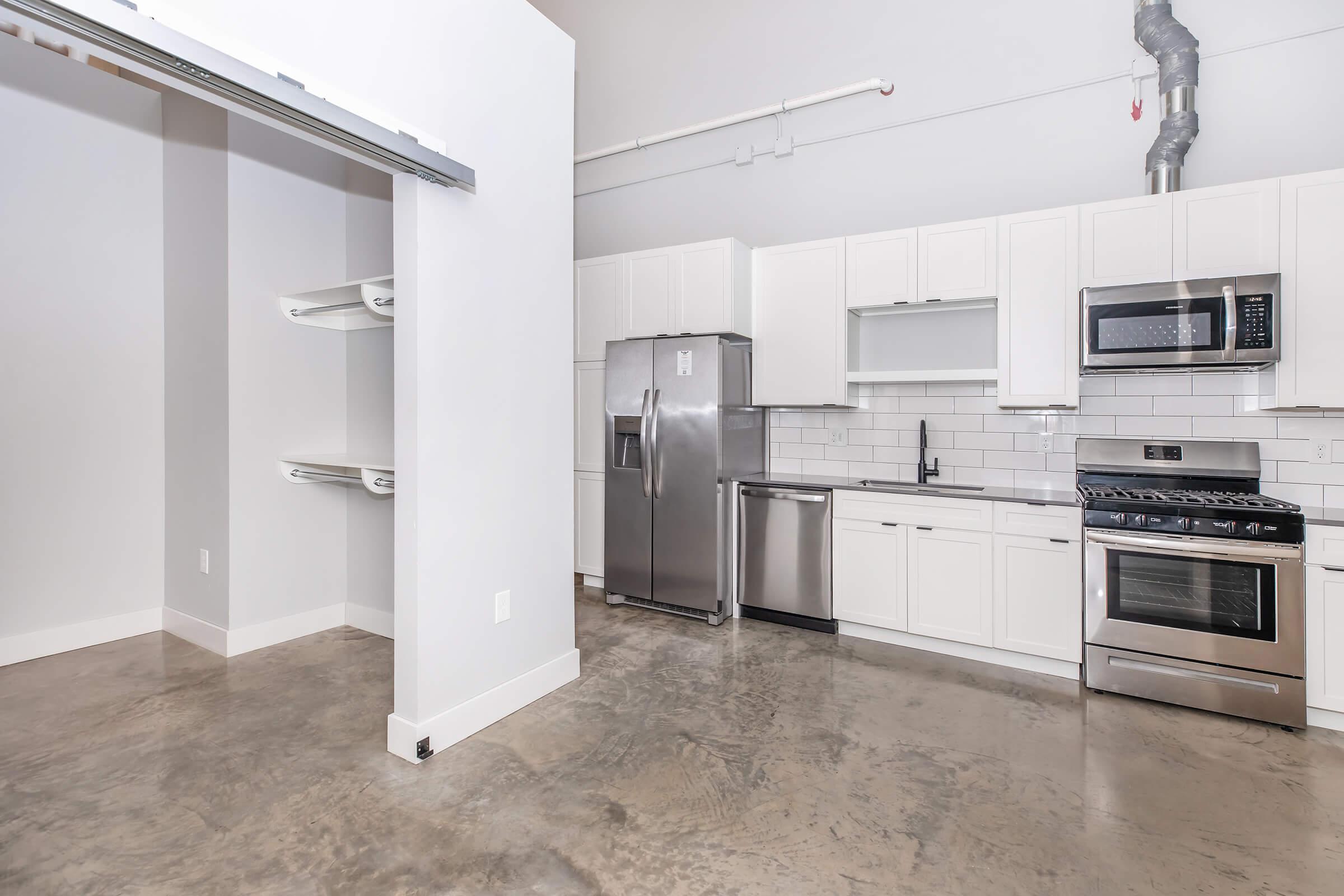 Modern kitchen featuring stainless steel appliances, including a refrigerator, microwave, and stove, along with white cabinetry. Concrete flooring enhances the contemporary aesthetic. An adjacent open space with shelves is visible, allowing for additional storage or design options.