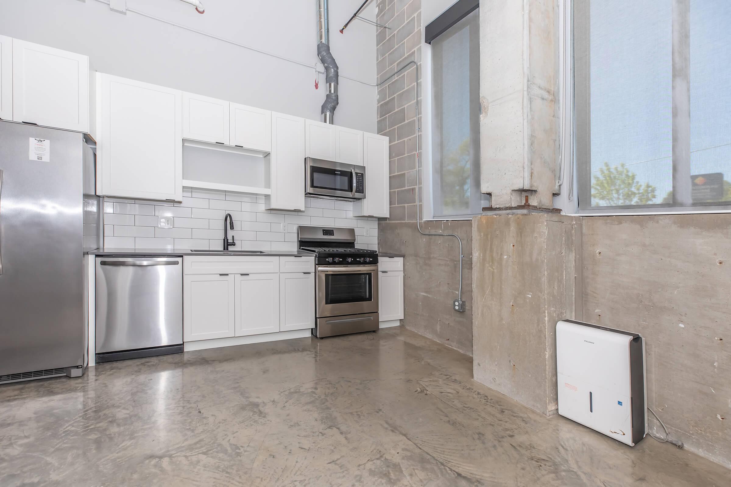 Modern kitchen in an industrial-style apartment featuring stainless steel appliances, including a refrigerator, microwave, and gas stove. The cabinetry is white with a sleek backsplash, while the flooring is polished concrete. Large windows provide natural light, and exposed concrete elements enhance the contemporary aesthetic.