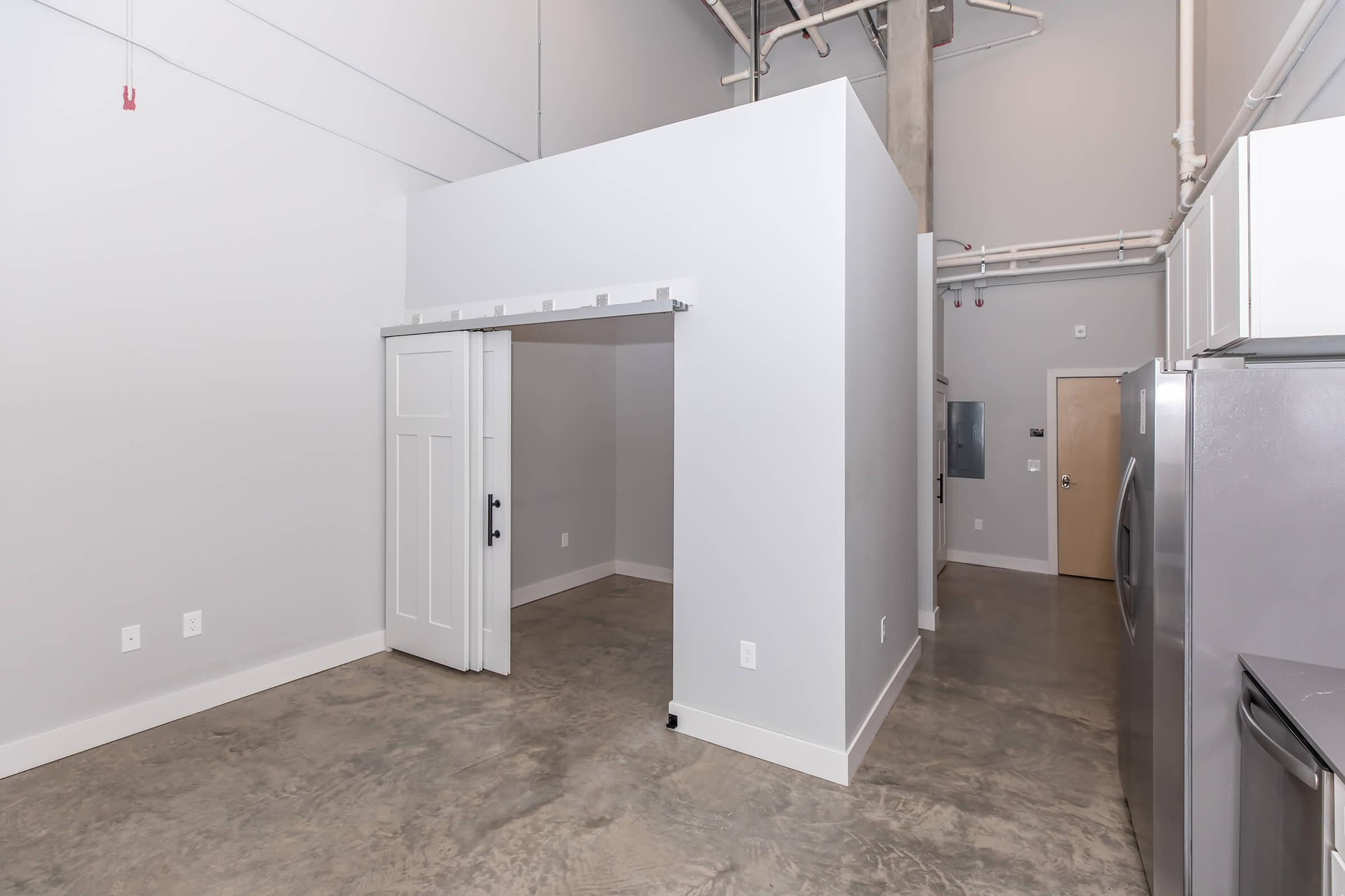 Interior view of a modern, minimalist space featuring gray walls and polished concrete floors. A small room with a white partition is visible, along with a door leading to another area. The ceiling has exposed pipes, and there are appliances seen in the background, contributing to an industrial aesthetic.
