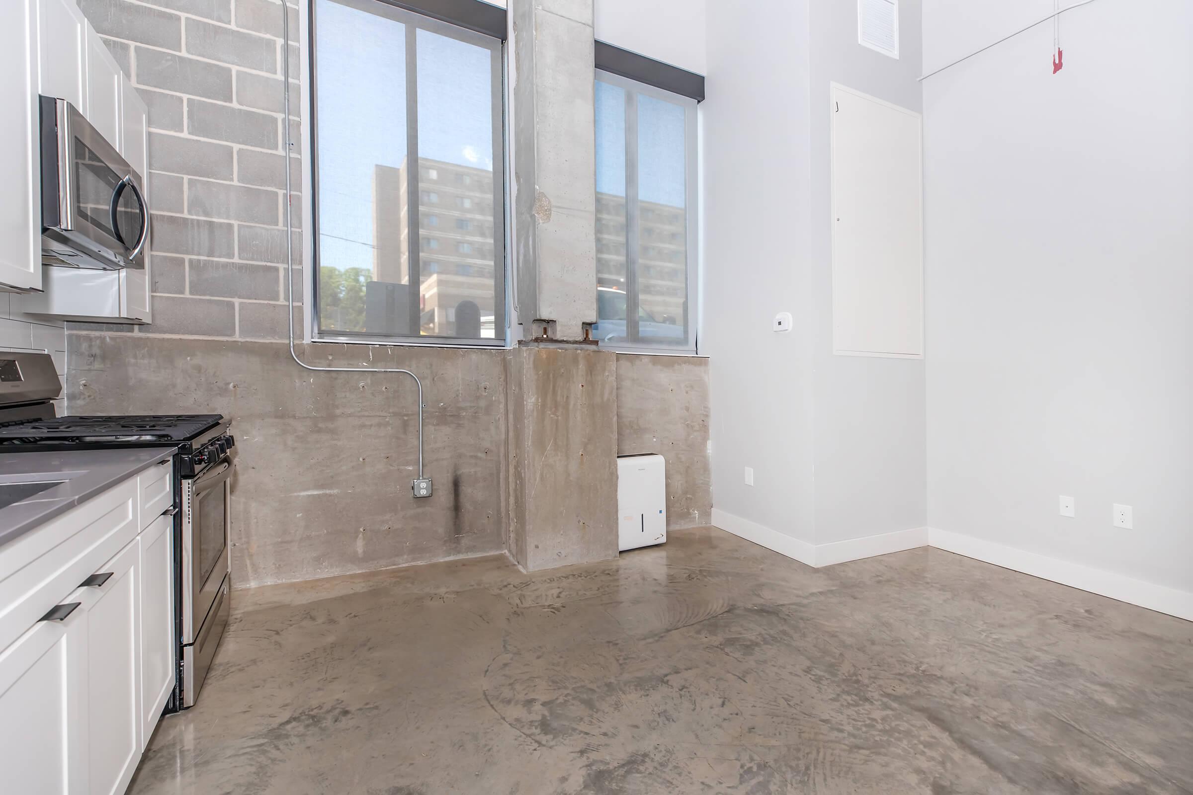 A modern apartment interior featuring a kitchen area with stainless steel appliances, including a gas stove, and a concrete wall. Large windows allow natural light, highlighting the polished concrete floor. The space has minimalist design elements and exposed industrial features, contributing to a contemporary aesthetic.