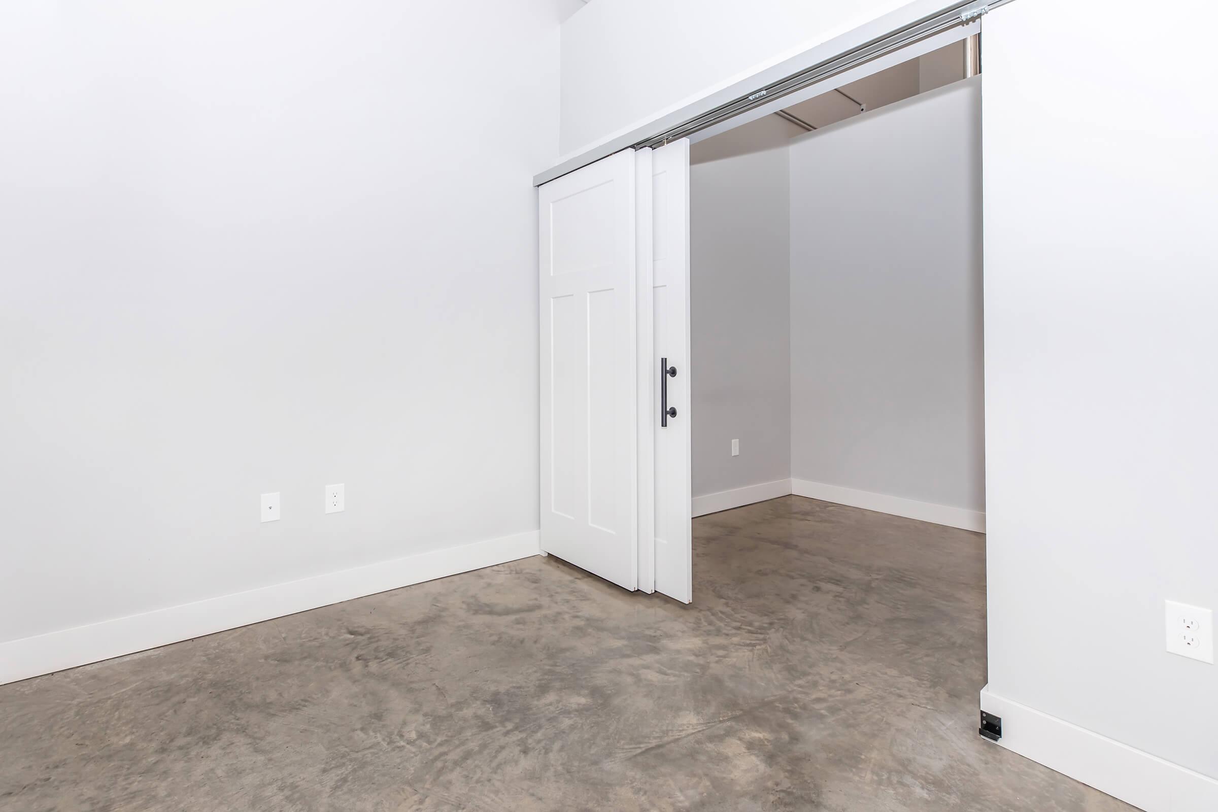 Interior view of a minimalist room featuring concrete floors, neutral gray walls, and a sliding door leading to another space. The room is empty, emphasizing a modern and spacious design, with no furniture or decorations present.