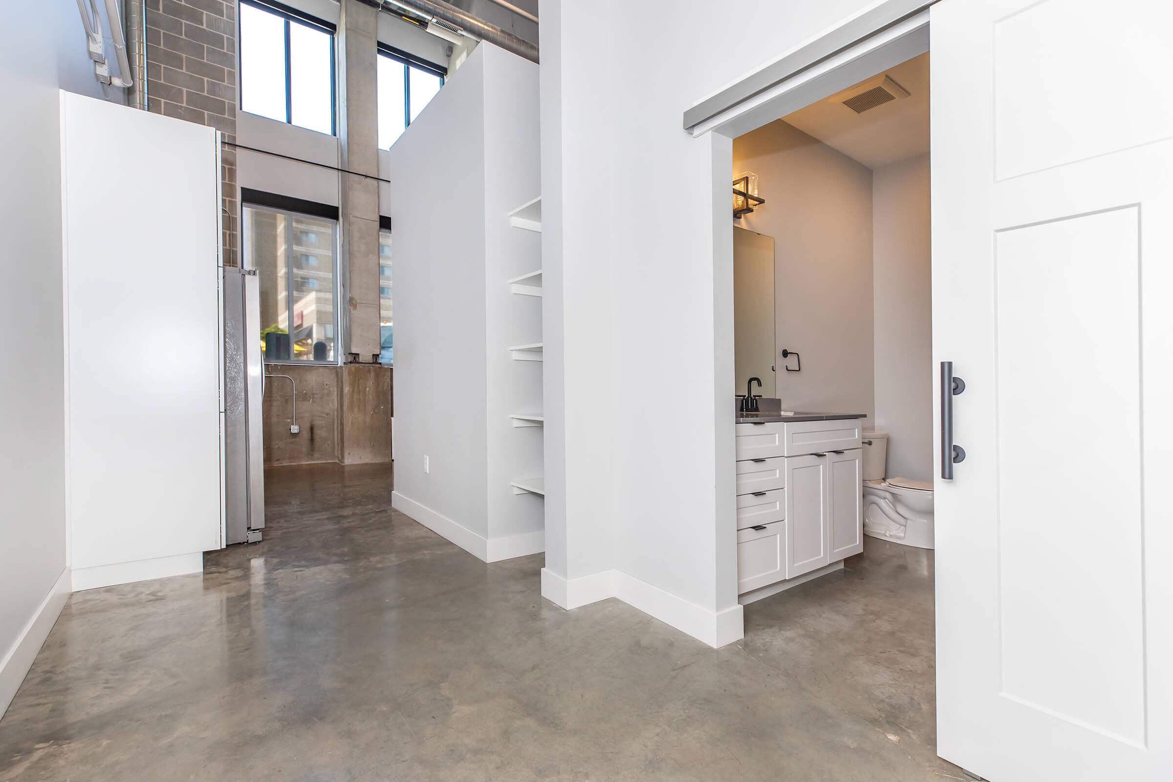 A spacious, modern interior featuring a polished concrete floor, white walls, and tall windows allowing natural light. On the left, a refrigerator is visible, while a bathroom is partially open on the right. The overall style is minimalist, with a clean and airy atmosphere.