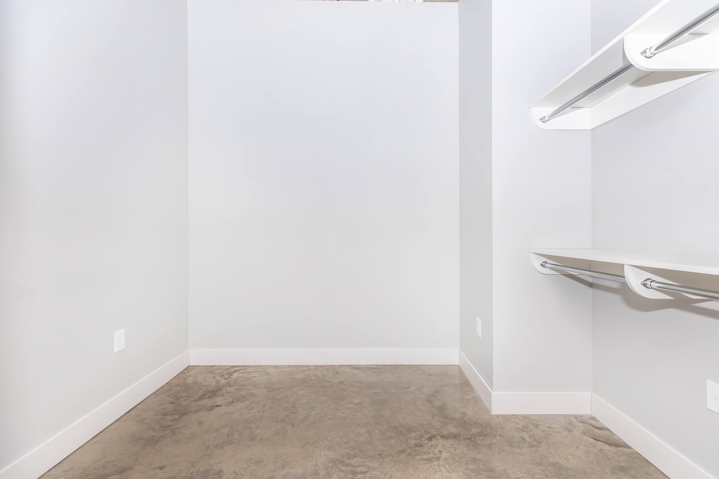 A spacious, empty closet featuring light gray walls, a concrete floor, and two open shelving units with metal rails on one side, providing ample storage and organization potential. The overall design emphasizes a minimalist aesthetic and functionality.