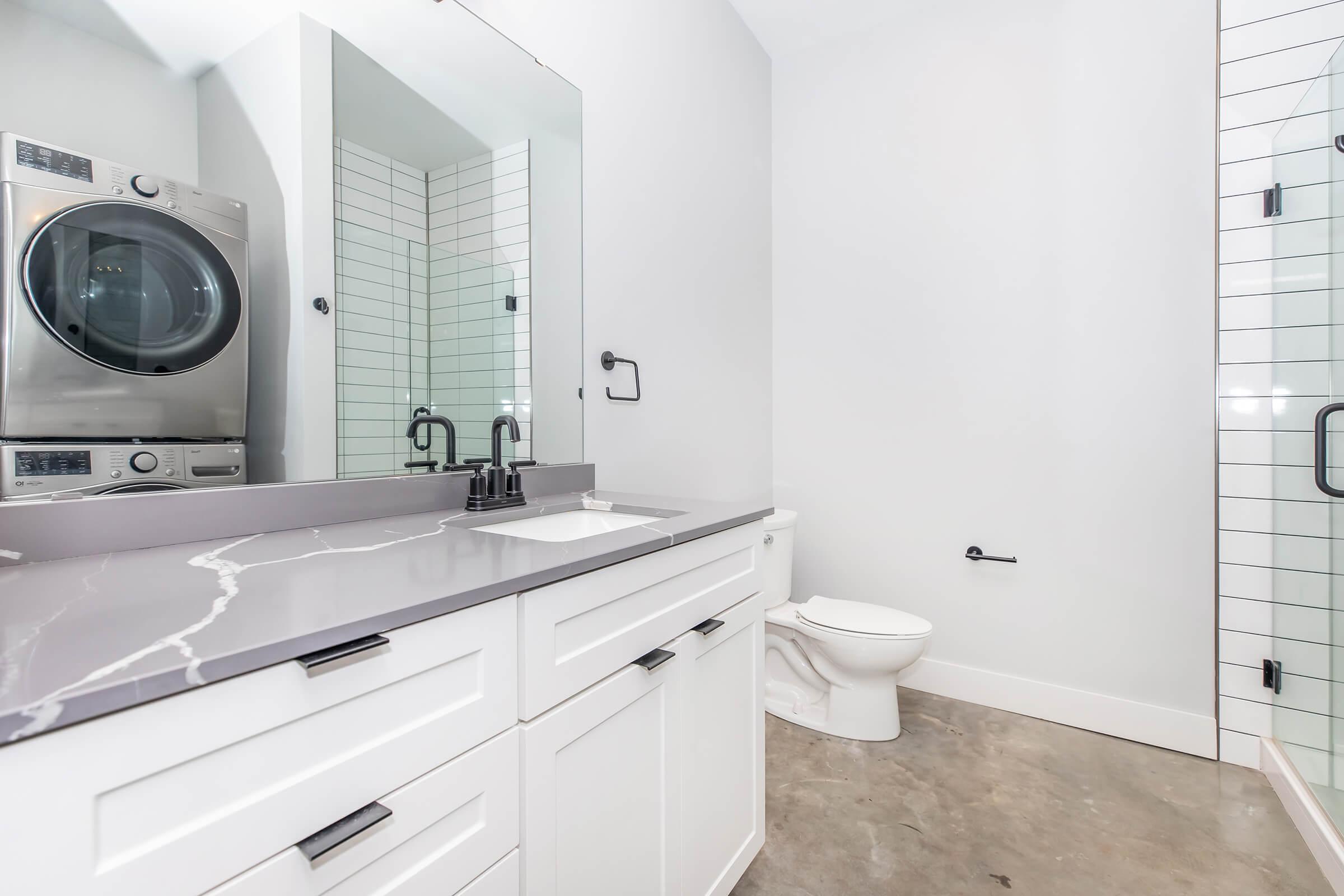 Modern bathroom featuring a sleek gray countertop with a sink, a large mirror, and a white cabinet below. To the left, there is a stacked washer and dryer unit. The space includes minimalist black fixtures, a glass shower enclosure, and a white toilet, with light-colored walls and concrete flooring.