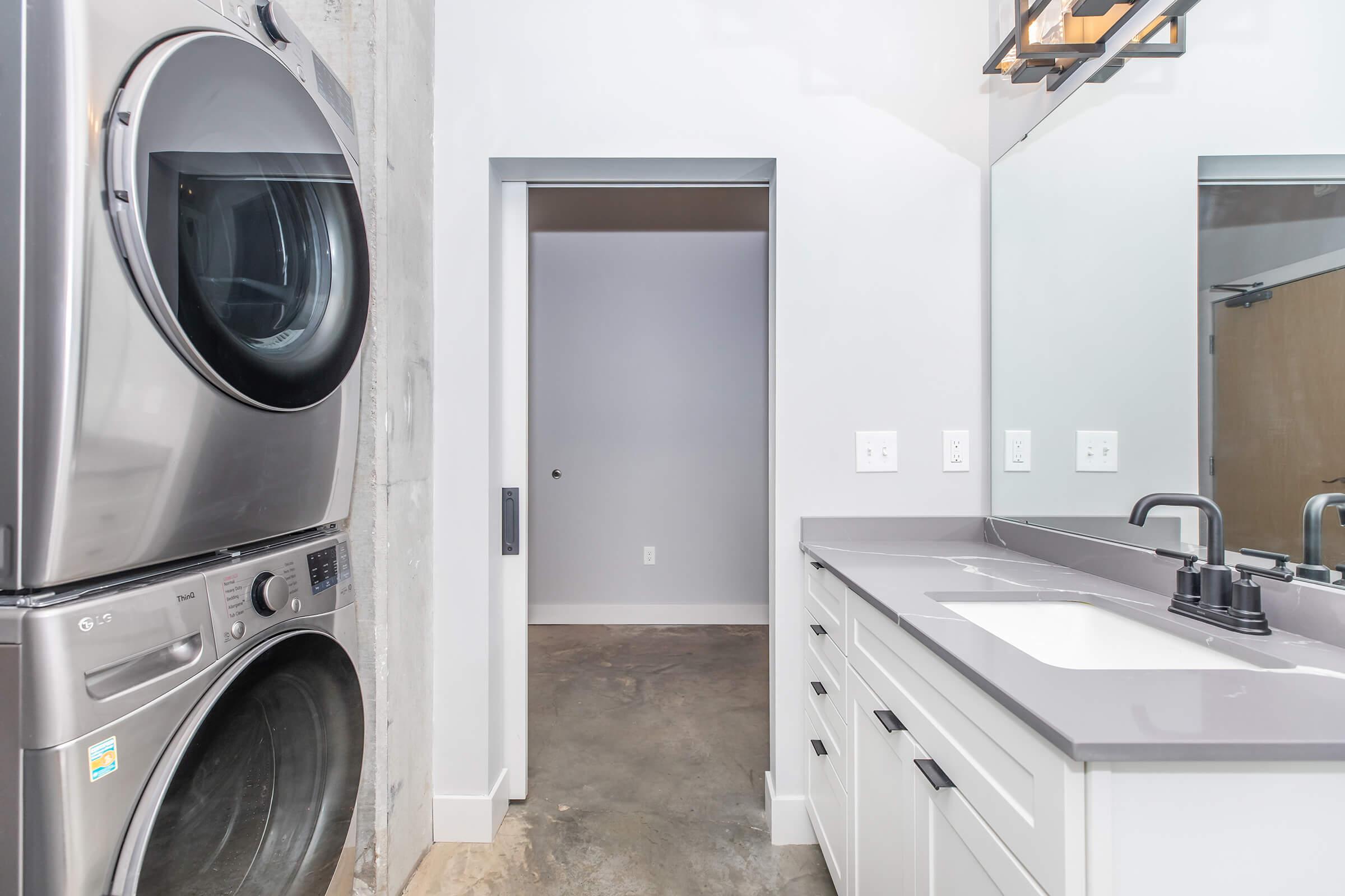 A modern laundry room featuring a stacked washer and dryer on the left, a sleek gray countertop with a black faucet and sink in the center, and a doorway leading to another room in the background. The walls are a mix of light colors and concrete, providing a contemporary aesthetic.