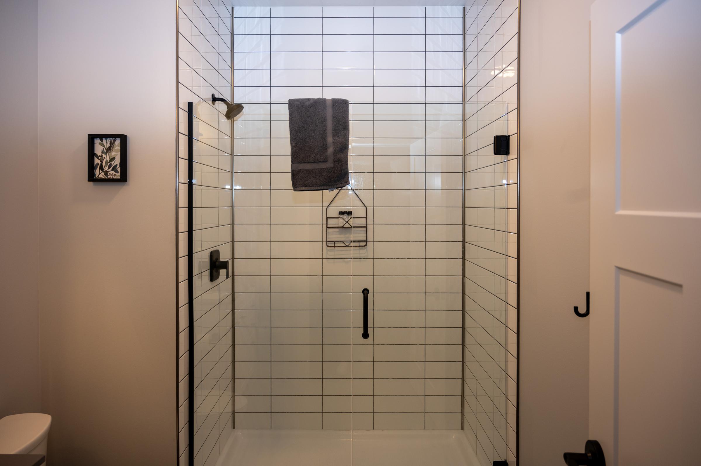 A modern shower space featuring white tiled walls, a glass door, a gray towel hanging on a rack, and black fixtures. A small framed artwork is displayed on the wall, and a white toilet is visible in the corner. The overall design is clean and minimalist.