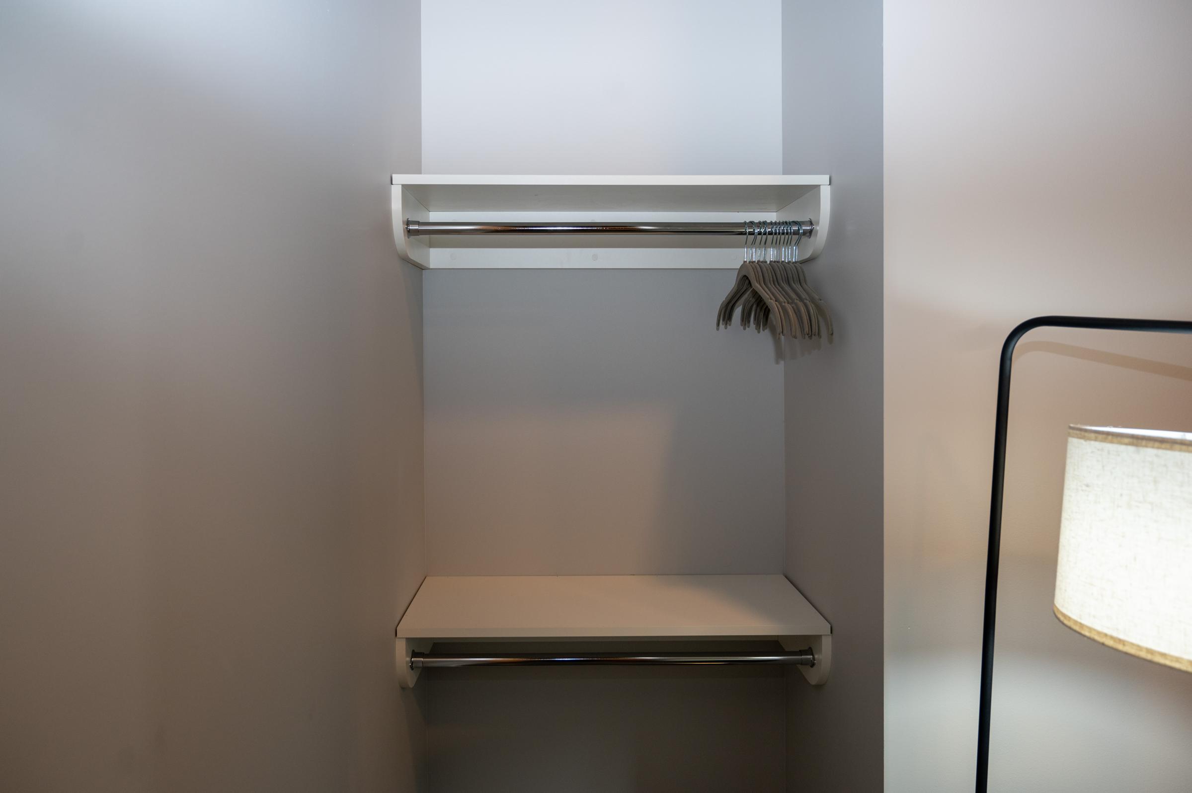 An empty closet featuring a white shelf and a chrome clothing rod with several hangers hanging from it. A second shelf is placed below the rod. A lamp is positioned nearby, casting soft light on the closet's interior. The walls are painted a light color, contributing to a clean and minimalistic appearance.