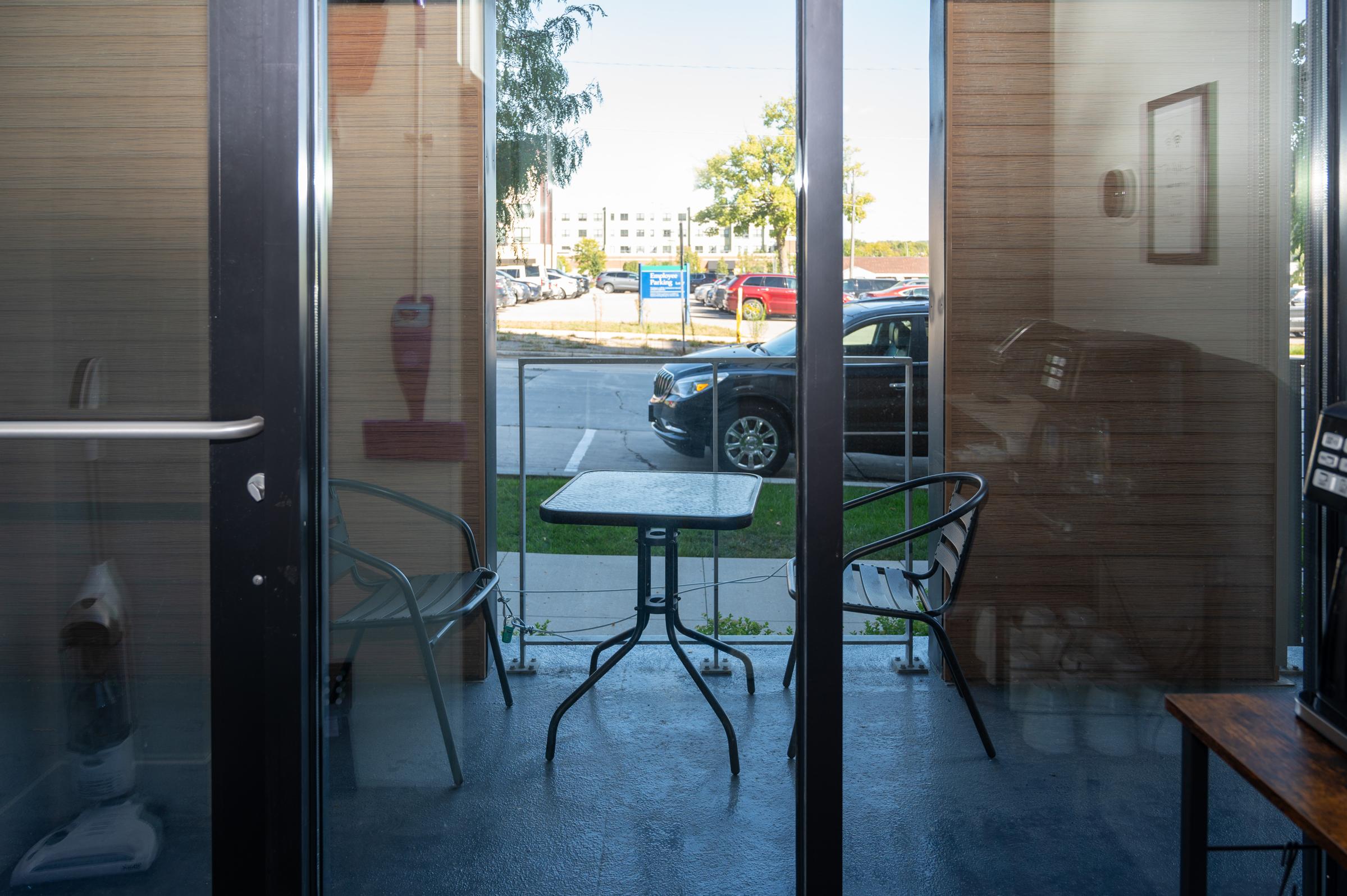 A small outdoor seating area visible through glass doors, featuring a table with two chairs. The background includes a parked car and landscaped greenery, providing a bright and inviting atmosphere.