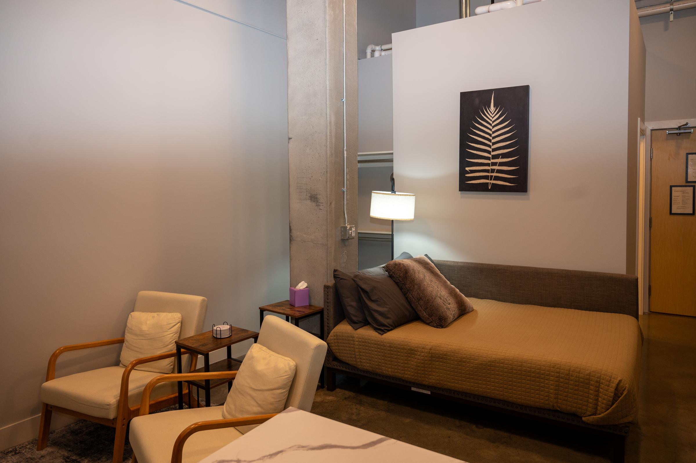 A cozy, modern bedroom featuring a queen-sized bed with decorative pillows, two armchairs, and a small side table. A wall art piece with a fern design hangs above the bed, while a floor lamp provides warm lighting. The walls are painted light gray, and the flooring is a polished concrete.