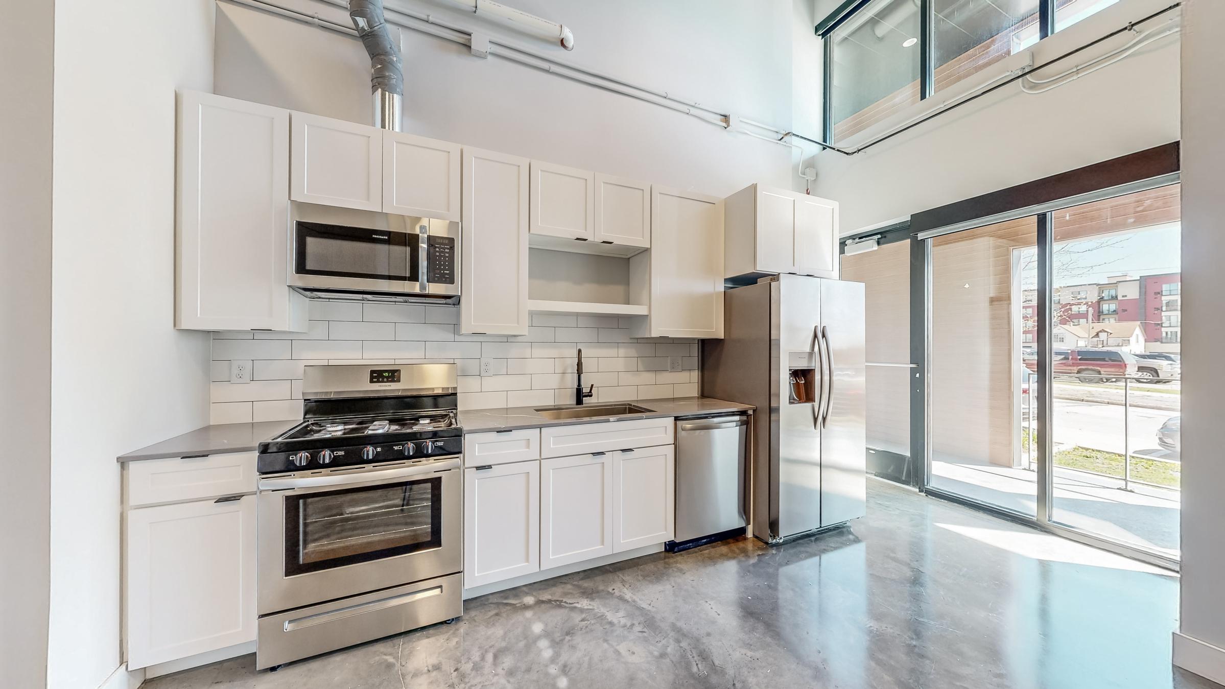 Modern kitchen featuring stainless steel appliances including a gas stove, microwave, and refrigerator. The white cabinetry contrasts with sleek countertops and a tiled backsplash. Large windows allow natural light to fill the space, highlighting the polished concrete floor and open layout.