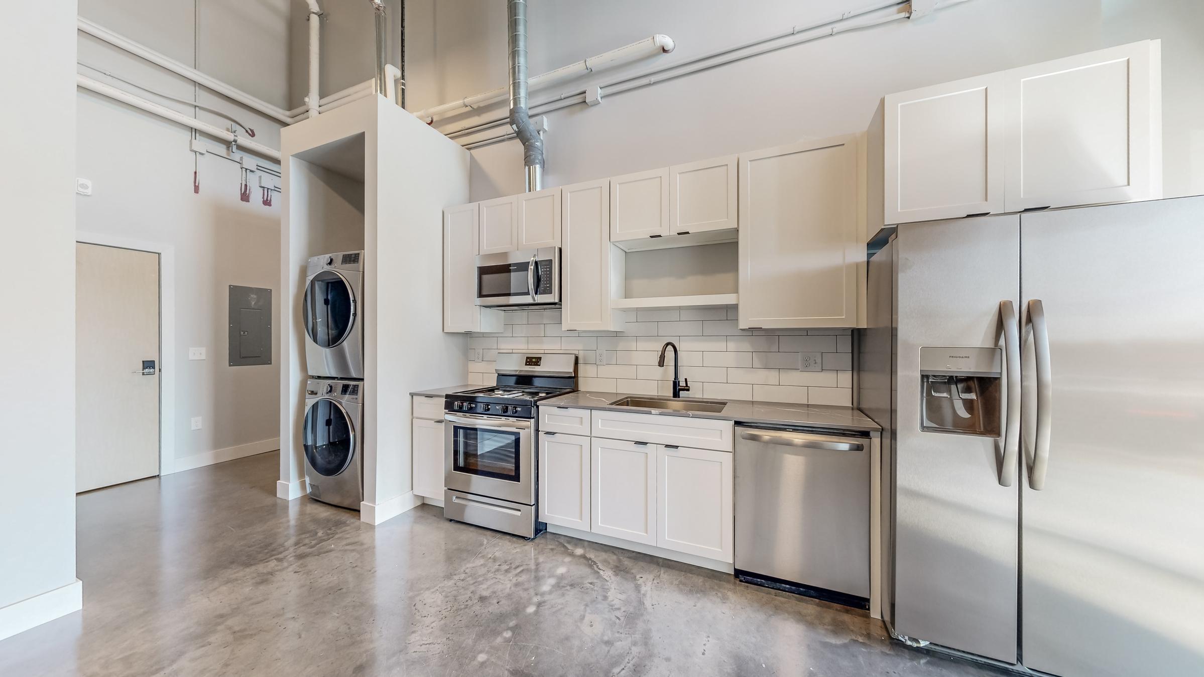 Modern kitchen featuring white cabinetry, stainless steel appliances including a refrigerator, stove, and dishwasher. A washer and dryer are stacked next to the kitchen area. The floor is polished concrete, and there are industrial-style pipes and vents visible on the ceiling.