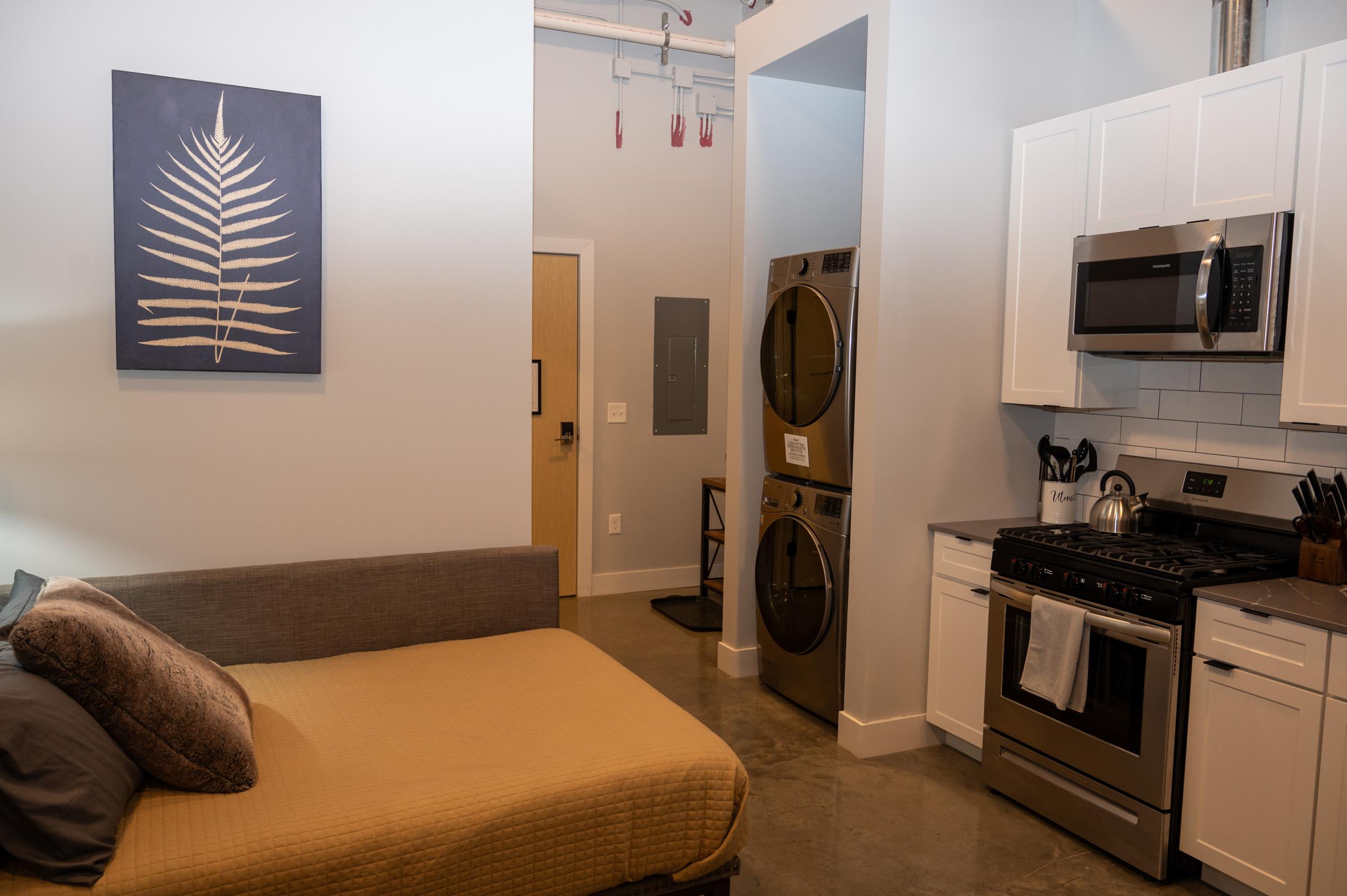 A modern studio apartment featuring a bed with a neutral-colored comforter, a kitchen area with stainless steel appliances, and a laundry nook with stacked washer and dryer. The walls are painted light colors, and there is a decorative fern print on the wall. The floor is polished concrete.