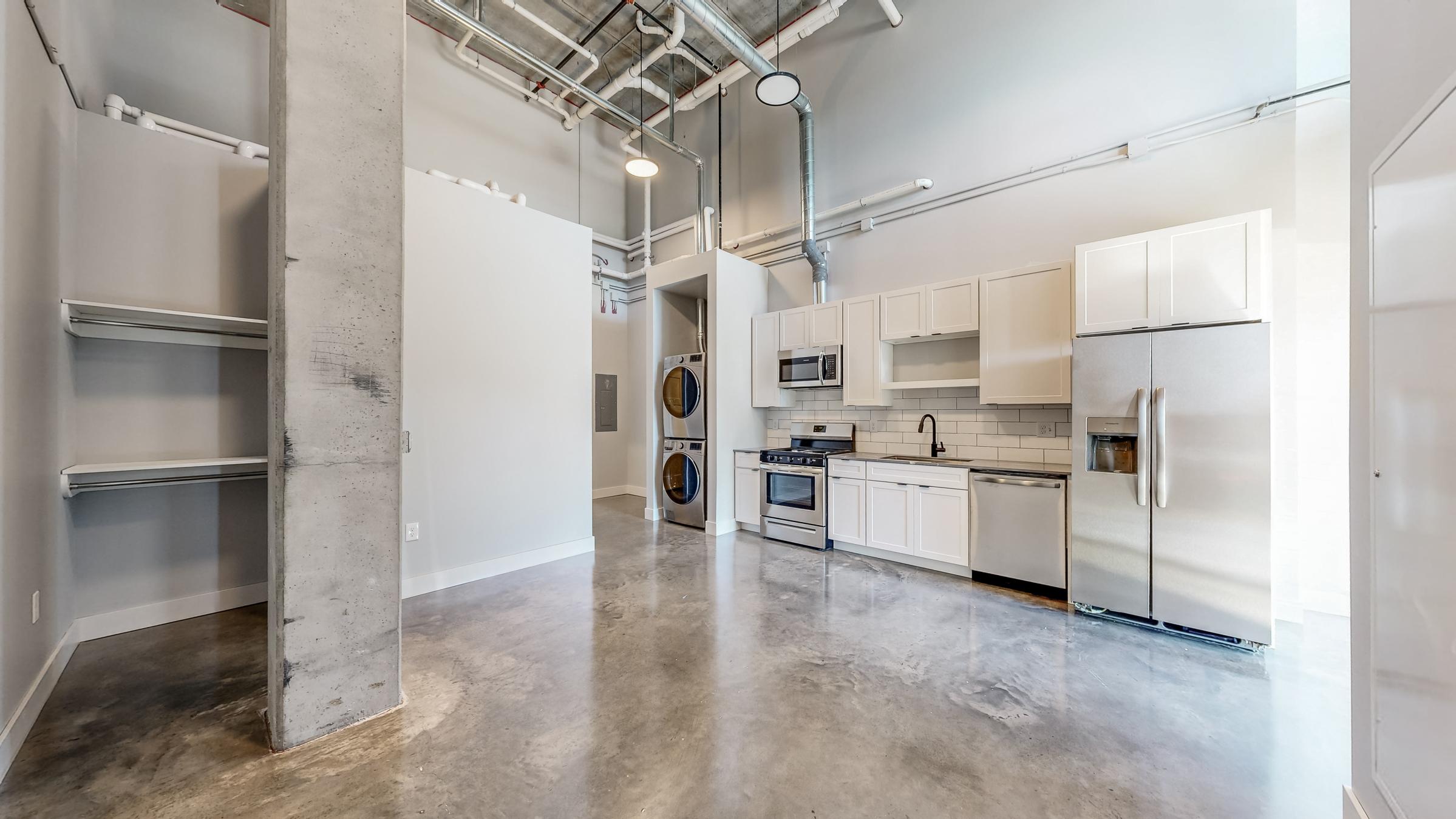 Modern kitchen in a spacious apartment, featuring stainless steel appliances, white cabinetry, and concrete floors. To the left, there are shelves and laundry machines. The open layout allows for ample natural light, complemented by high ceilings and industrial elements.