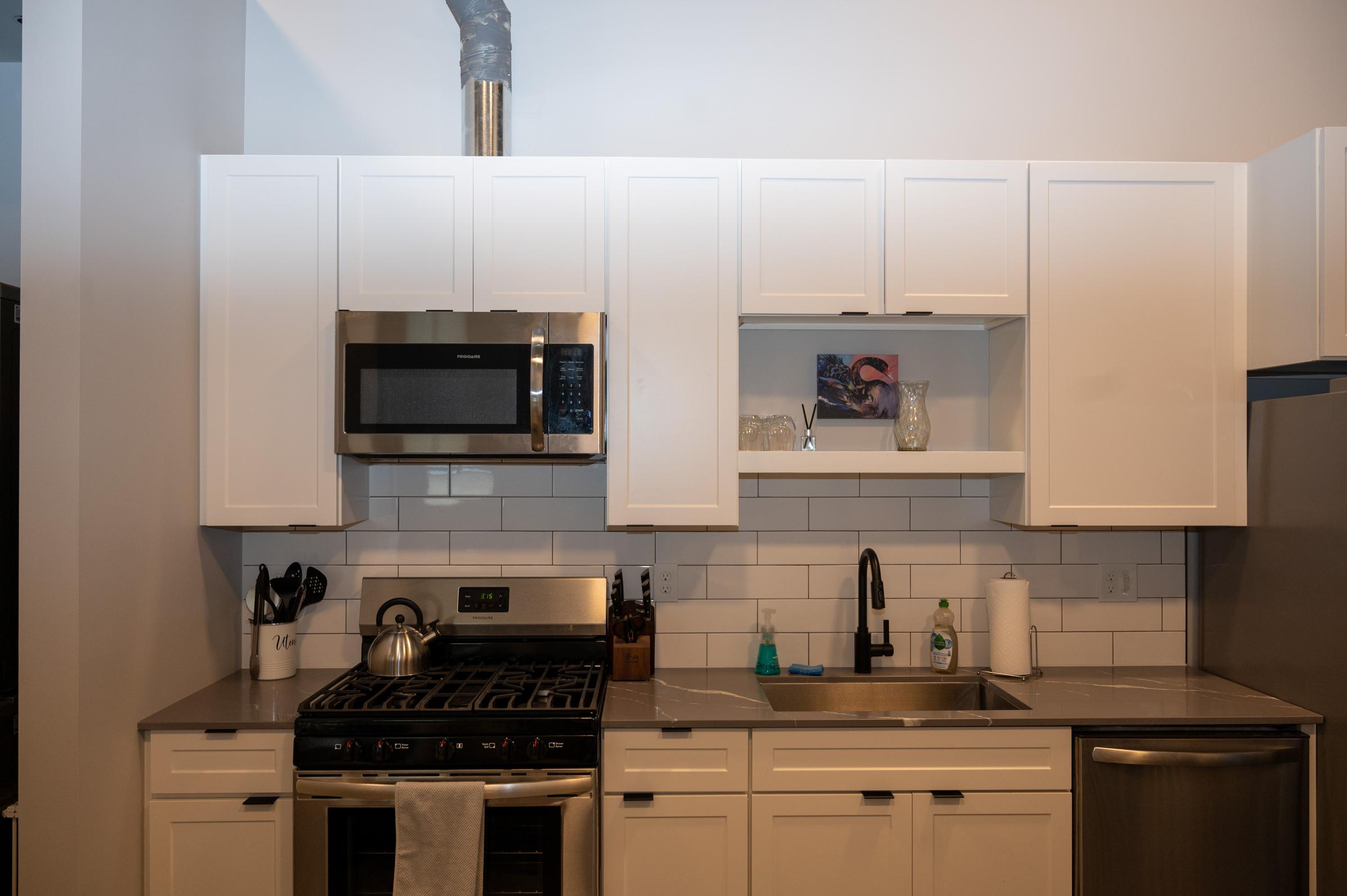 Modern kitchen featuring white cabinetry, a stainless steel microwave, and a gas stove. The countertop is gray with a sink and faucet. A few kitchen utensils are visible on the counter, along with dish soap and a decorative item in the open shelf. Natural light illuminates the space.