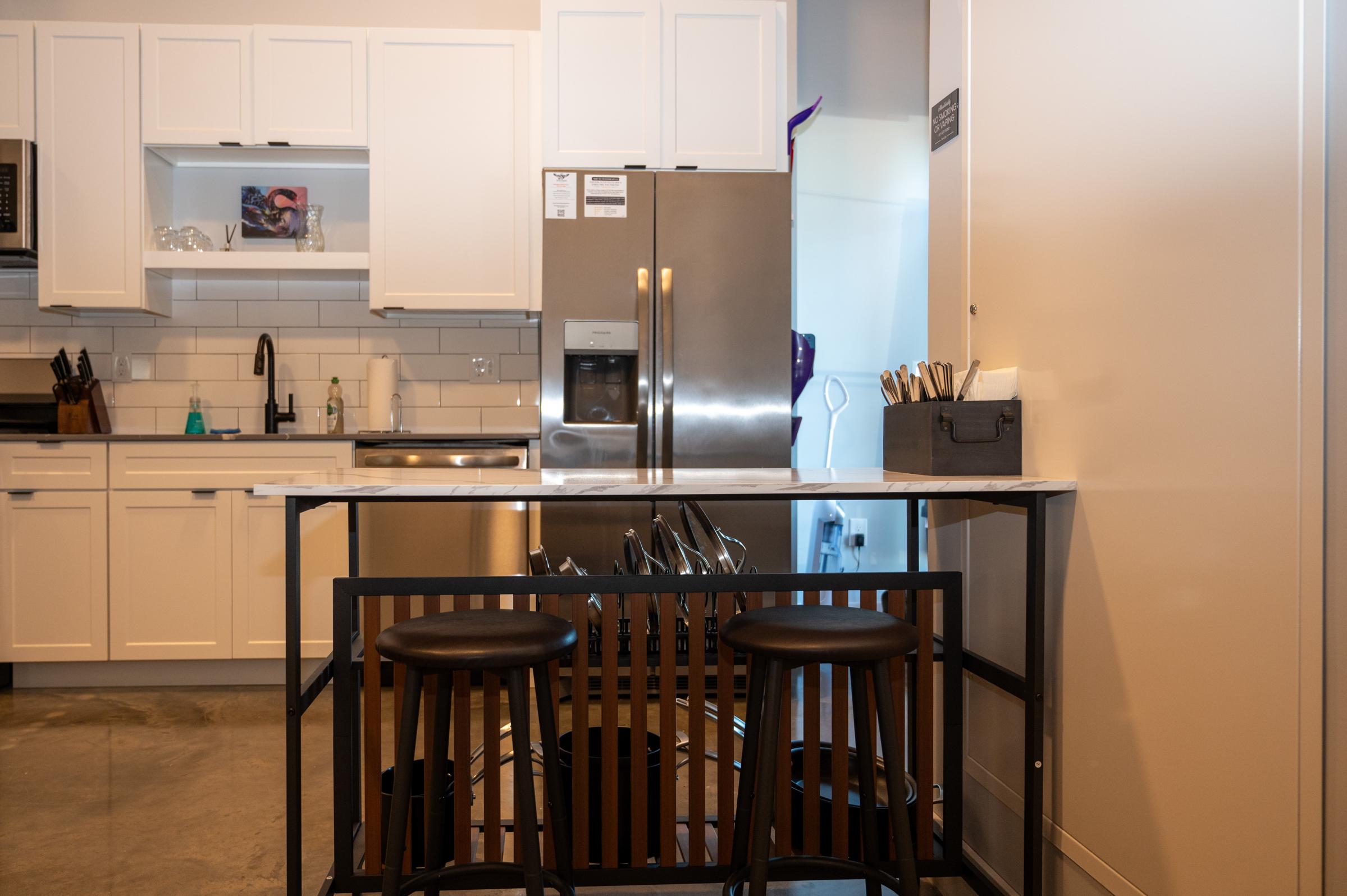 Modern kitchen with white cabinetry and stainless steel appliances. A marble-topped island with three black stools is in the foreground. Kitchen tools and utensils are organized in a small caddy on the island, while a silver refrigerator and a window are visible in the background.
