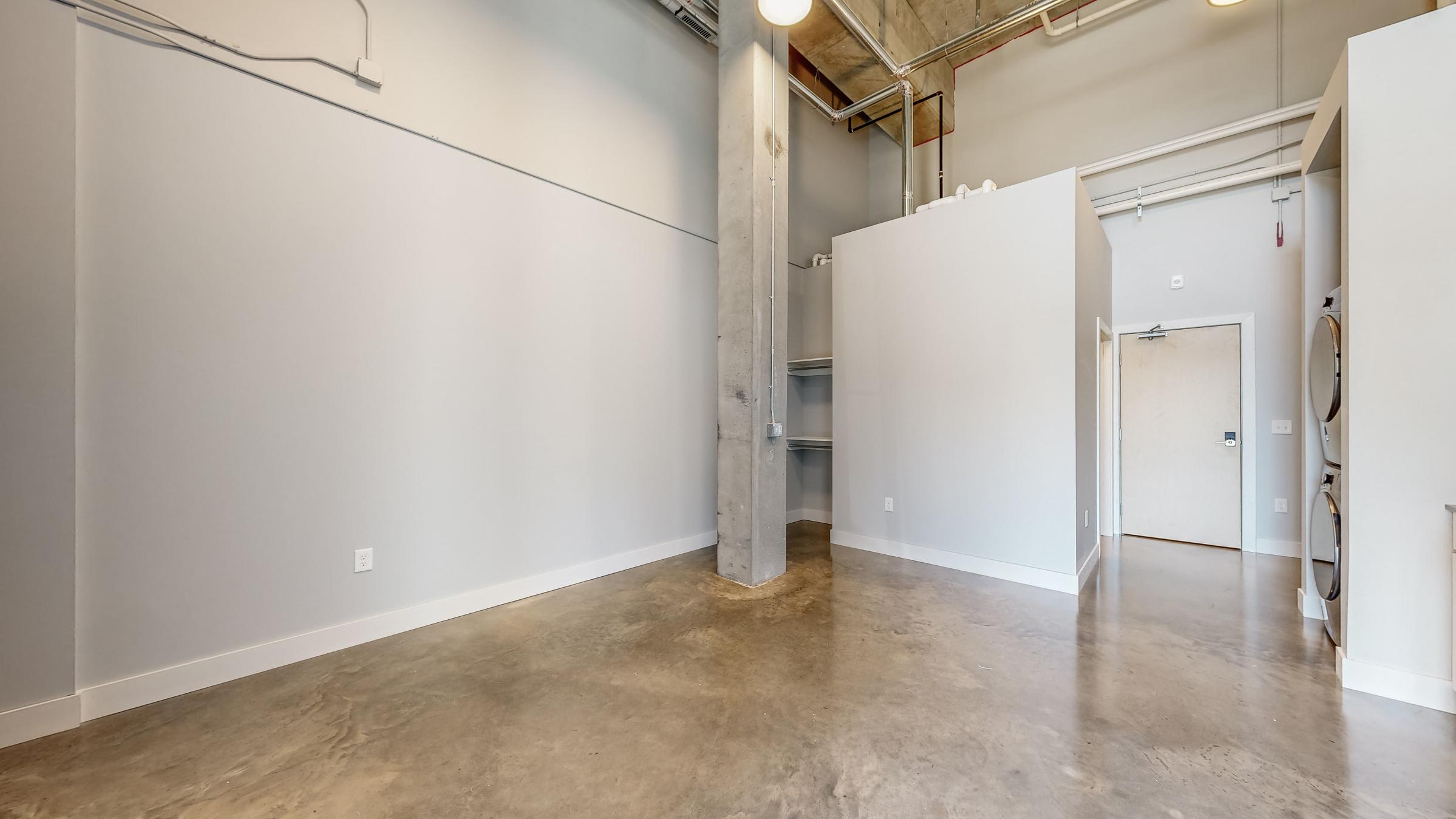 A spacious, minimalist room with concrete flooring and gray walls. On the left, there is a concrete pillar, and to the right, a white storage closet with a door is visible. Natural light enters the space from above, and the area feels open and modern, suitable for various uses.