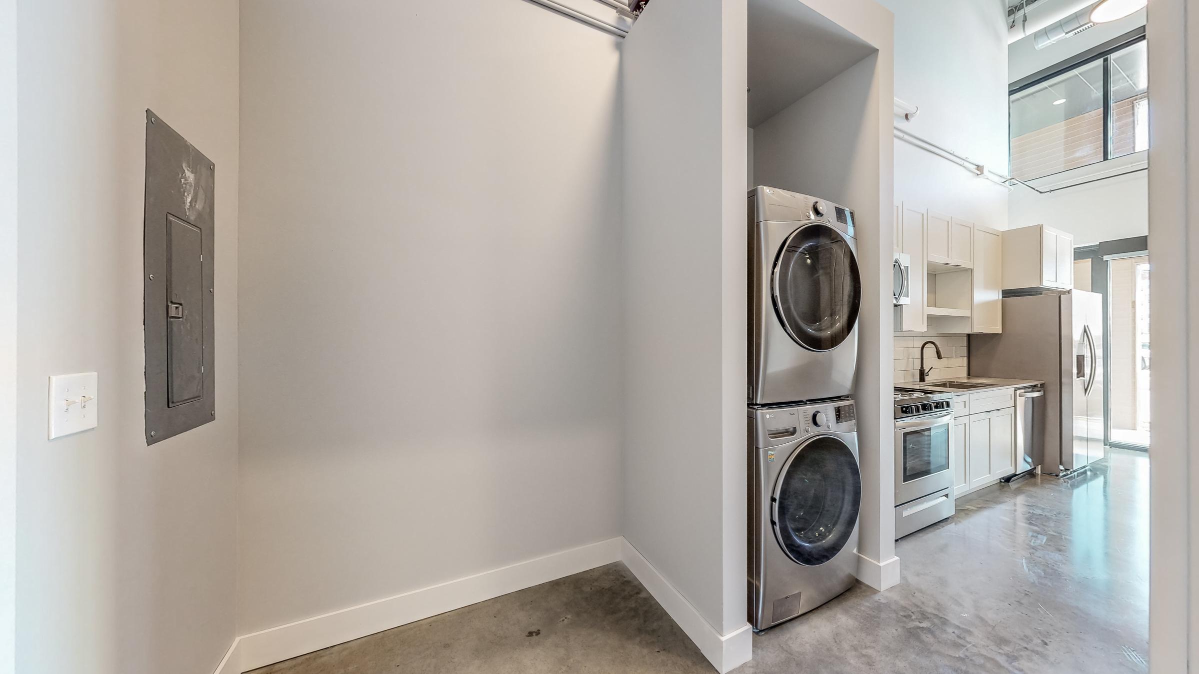 A modern laundry area featuring a stacked washer and dryer unit next to a kitchen with stainless steel appliances. The room has smooth concrete floors, light gray walls, and large windows providing natural light. The layout is open and spacious, contributing to a contemporary design.