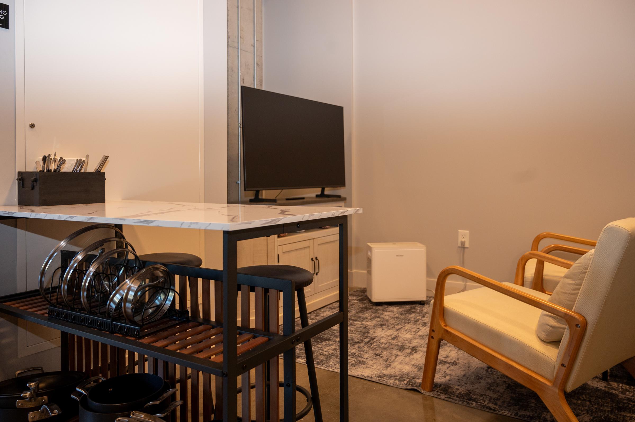 A cozy interior space featuring a small dining area with a marble-top table and bar stools, a TV on a stand, an armchair, and a white mini fridge. The flooring is polished concrete, complemented by a rug, and there are kitchen utensils in a black container on the table. The walls are light gray.