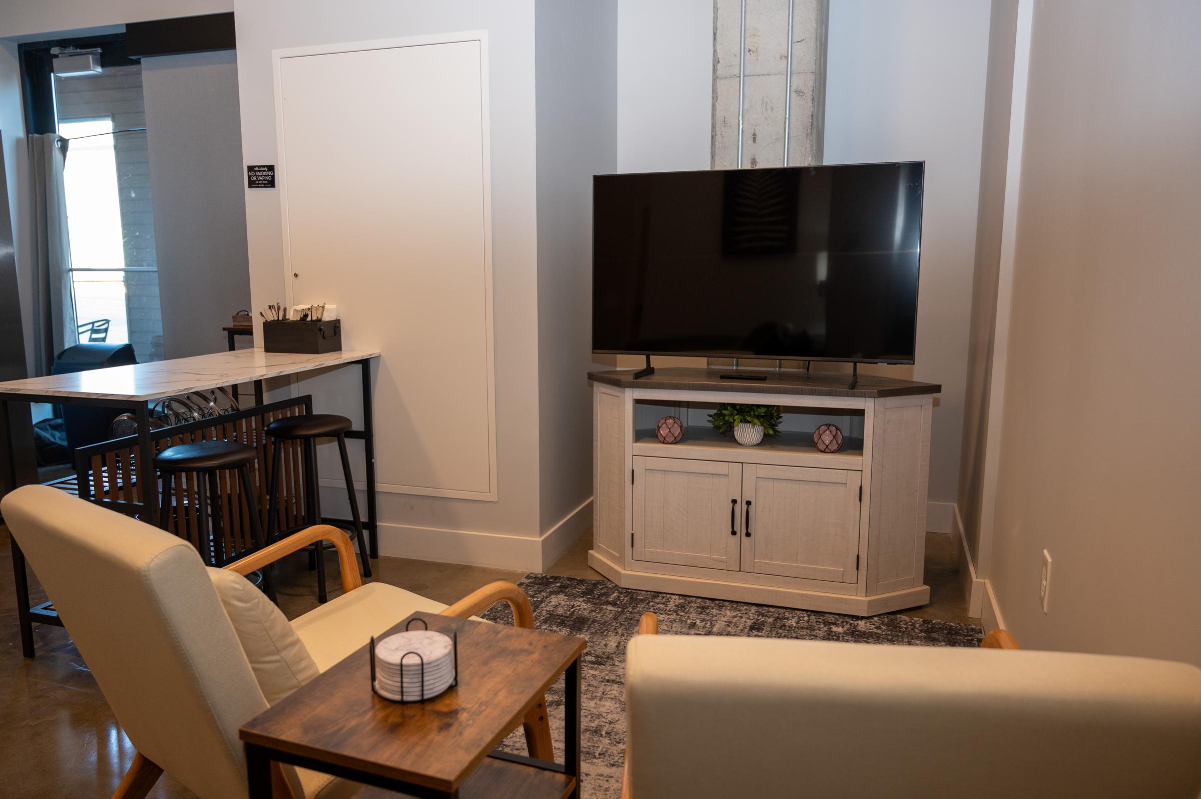A cozy living space featuring a modern television on a white cabinet, two light-colored armchairs, a wooden side table with a decorative candle, and a bar area with stools. The room has a neutral color palette with a rug and natural light illuminating the space.
