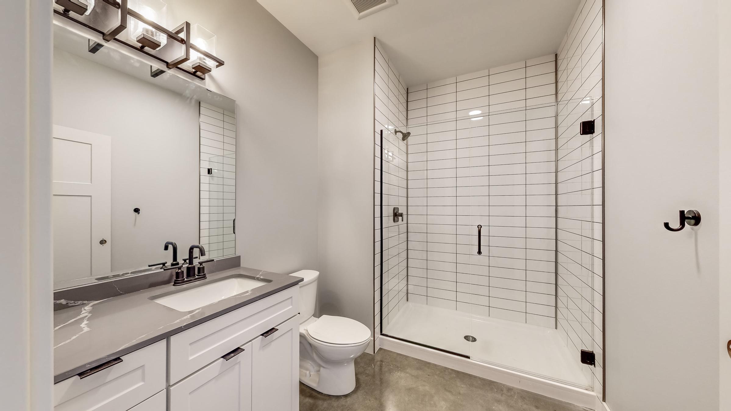 Modern bathroom featuring a sleek design with a gray countertop sink, white cabinetry, a standing shower with black fixtures, and large white tiles. The mirror above the sink reflects the bright, neutral walls and lighting fixtures, while a toilet is positioned beside the sink area.