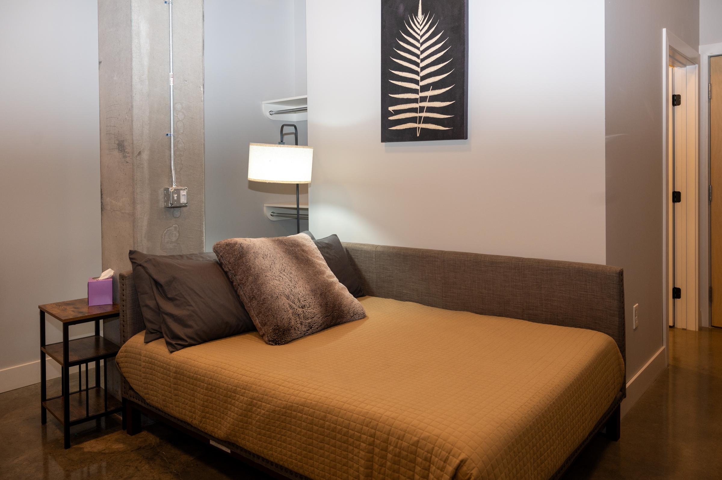 Cozy bedroom with a beige quilted bedspread and gray pillows. A small bedside table holds a lamp, and a piece of wall art featuring a leaf is displayed. The room has a modern industrial feel with exposed concrete and light-colored walls.
