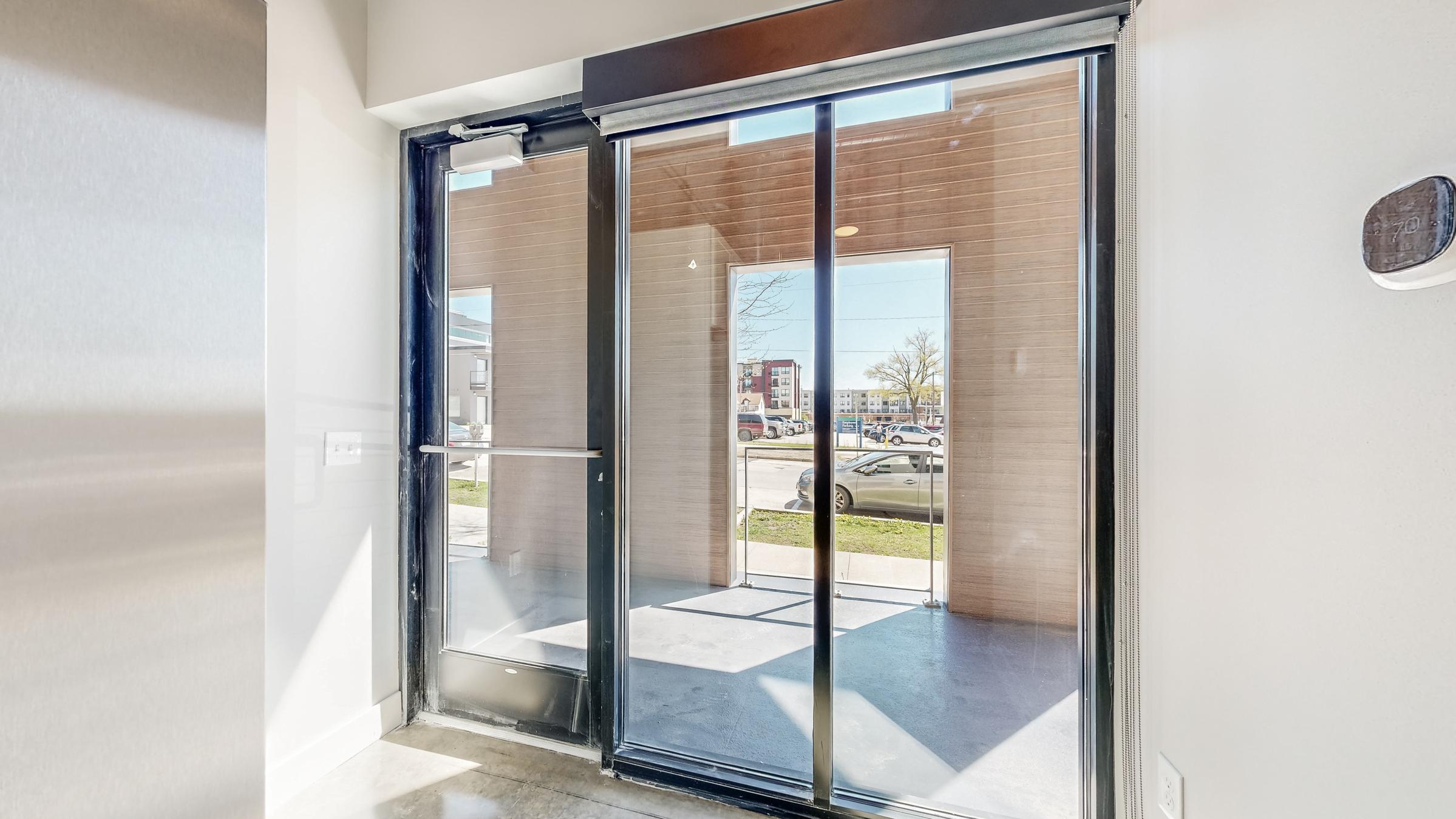 A modern entrance with sliding glass doors leading to an outdoor area. The interior features polished concrete flooring and a neutral-colored wall. Outside, vehicles are parked along a road, with a glimpse of buildings and trees in the background. Natural light illuminates the space.