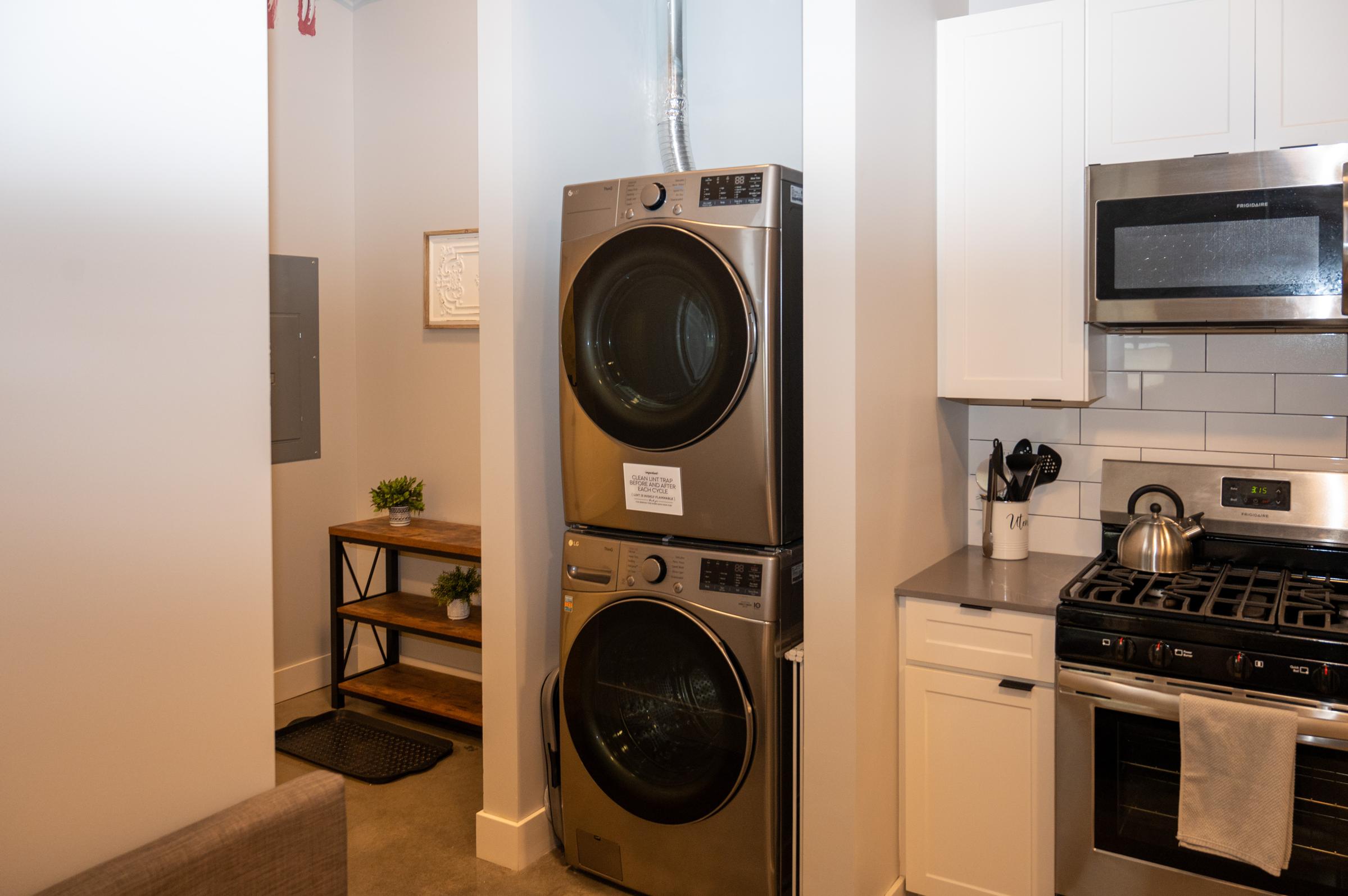 A modern laundry area featuring stacked stainless steel washer and dryer units next to a small wooden shelf with plants. The adjacent kitchen includes a stainless steel stove, microwave, and light-colored cabinetry against a tiled backsplash, creating a stylish, functional space.