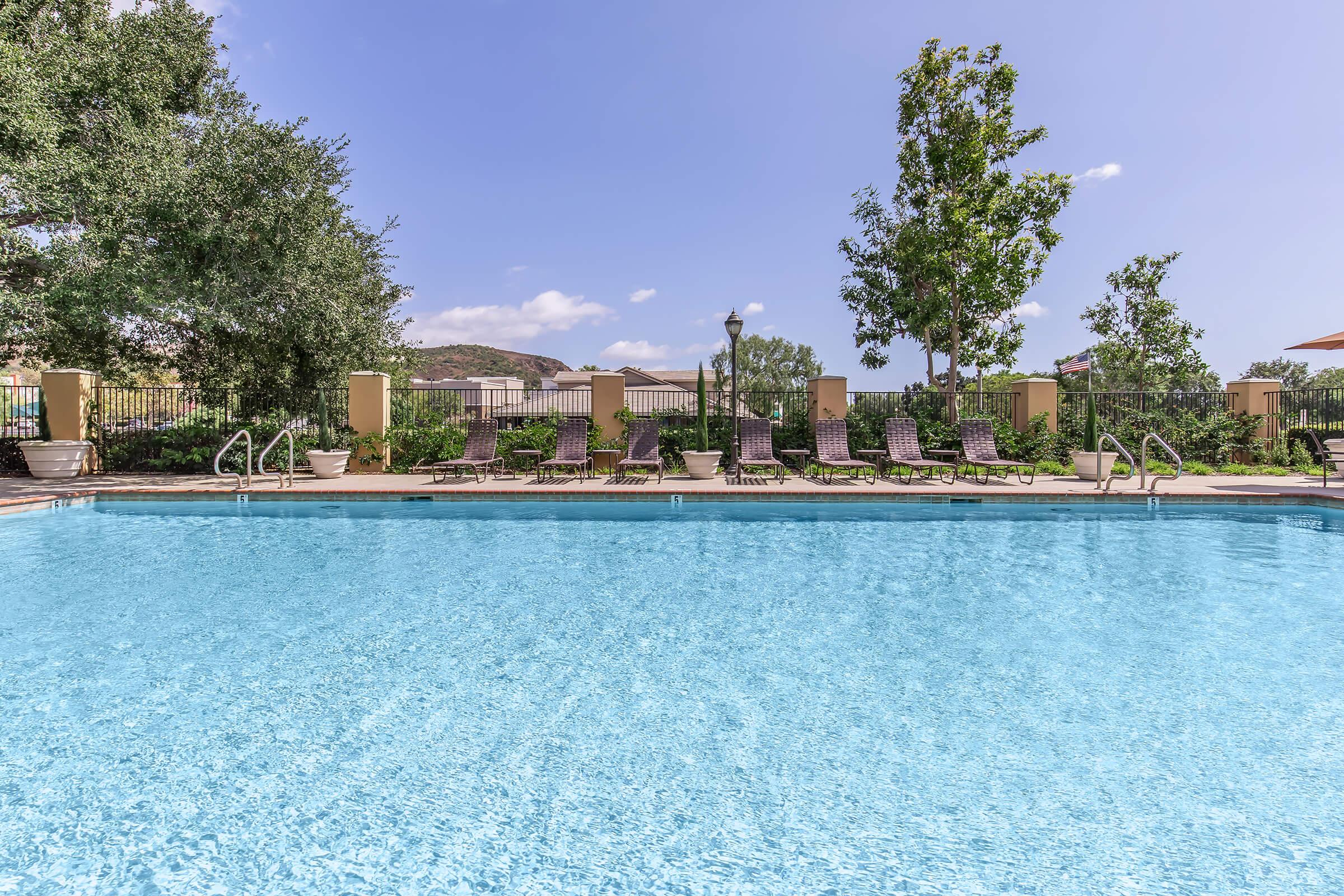 A clear blue swimming pool surrounded by lounge chairs. Lush greenery and trees line the area, and there are umbrellas for shade. The sky is bright with a few clouds, creating a serene outdoor space.