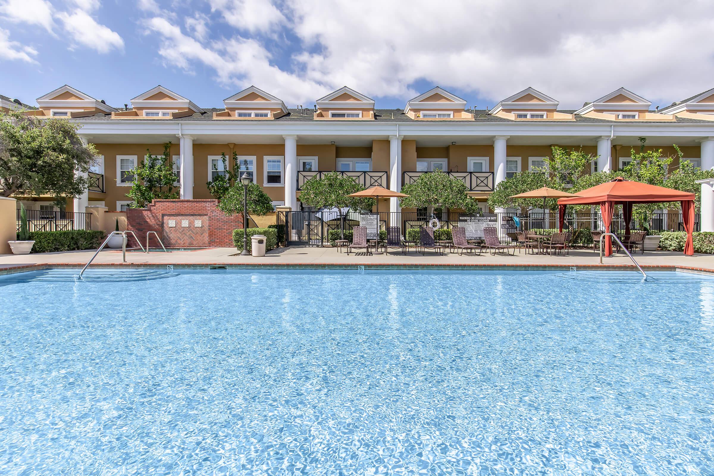 A clear blue pool surrounded by lounge chairs and tables, with an orange canopy providing shade. In the background, two-story buildings with columns and balconies are visible, along with well-maintained trees and landscaping. The sky is partly cloudy, creating a bright and inviting atmosphere.
