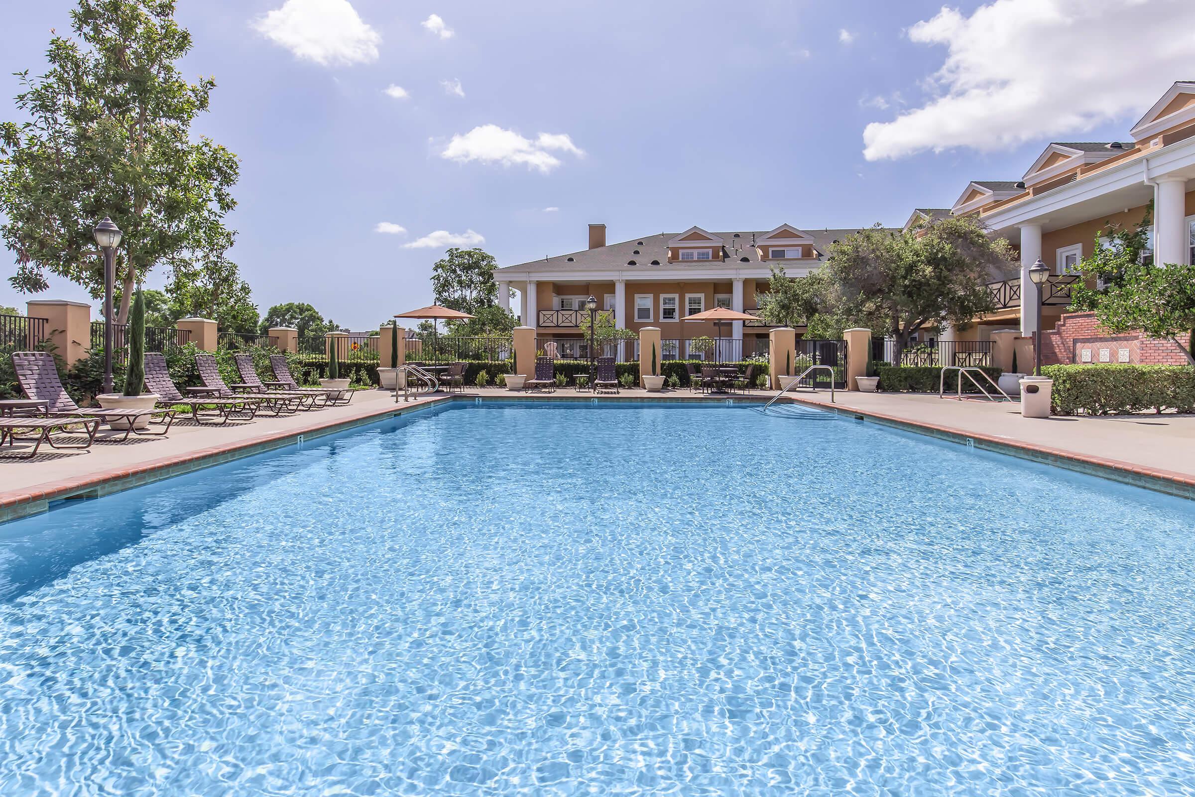 A spacious outdoor swimming pool surrounded by lounge chairs and umbrellas, with a well-maintained landscaped area in the background. Modern apartment buildings are visible beyond the pool, under a clear blue sky with a few fluffy clouds.