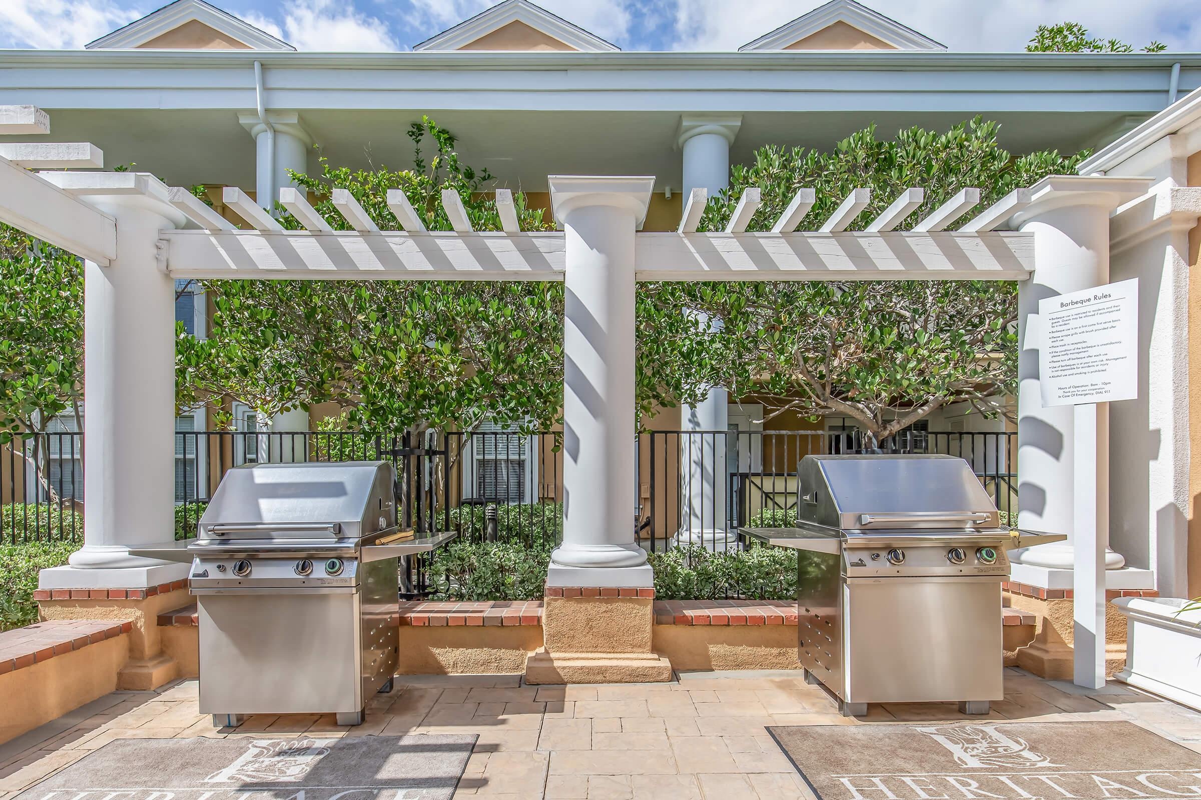 Two stainless steel outdoor grills located under a pergola in a landscaped area, surrounded by greenery. The scene features white columns and a stone pathway, contributing to a welcoming atmosphere. A sign is visible in the background, providing additional information.