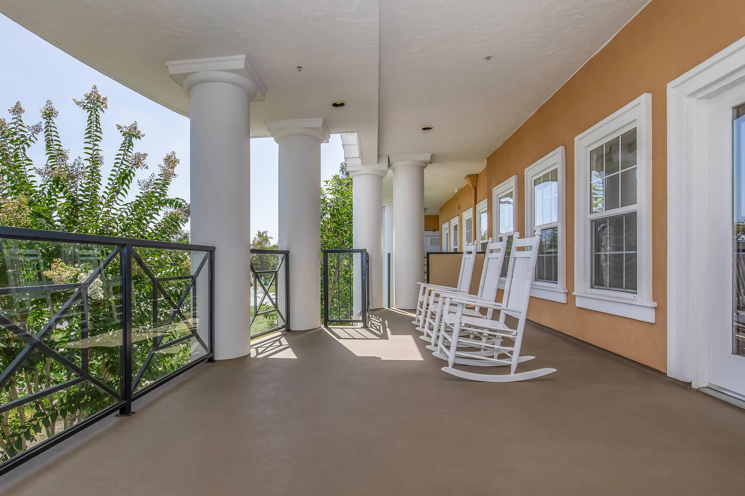 A spacious porch featuring white rocking chairs and decorative pillars. The porch overlooks greenery and is illuminated by natural light, creating a welcoming atmosphere. The walls are painted in warm tones, enhancing the inviting scene.