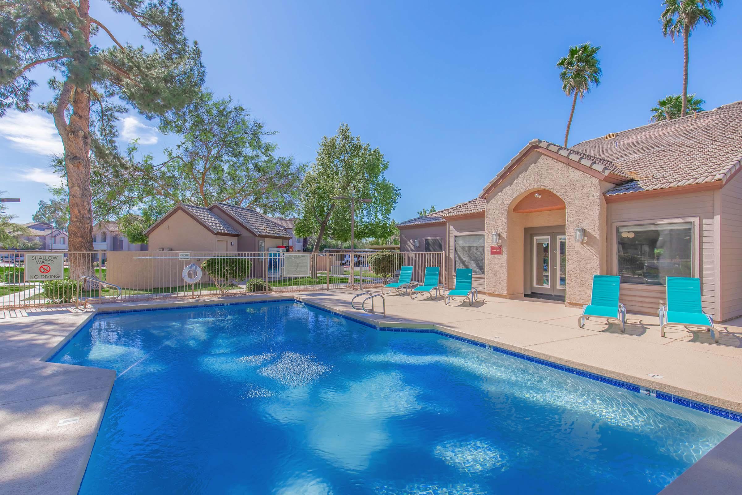 A clear blue swimming pool surrounded by lounge chairs and palm trees, with a well-maintained garden area and residential buildings in the background. The sky is bright and sunny, creating a cheerful and inviting atmosphere.