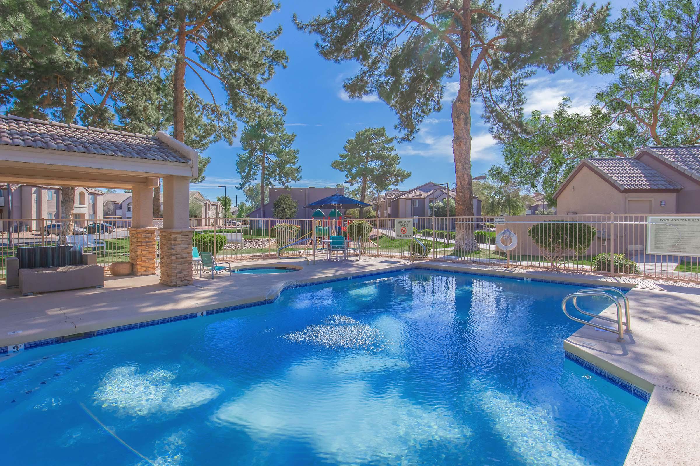 A clear blue swimming pool with a gentle slope leading into it, surrounded by green trees and residential buildings. Sunlight casts reflections on the water's surface, creating a tranquil and inviting atmosphere. Lounge chairs and a shaded area are visible nearby.