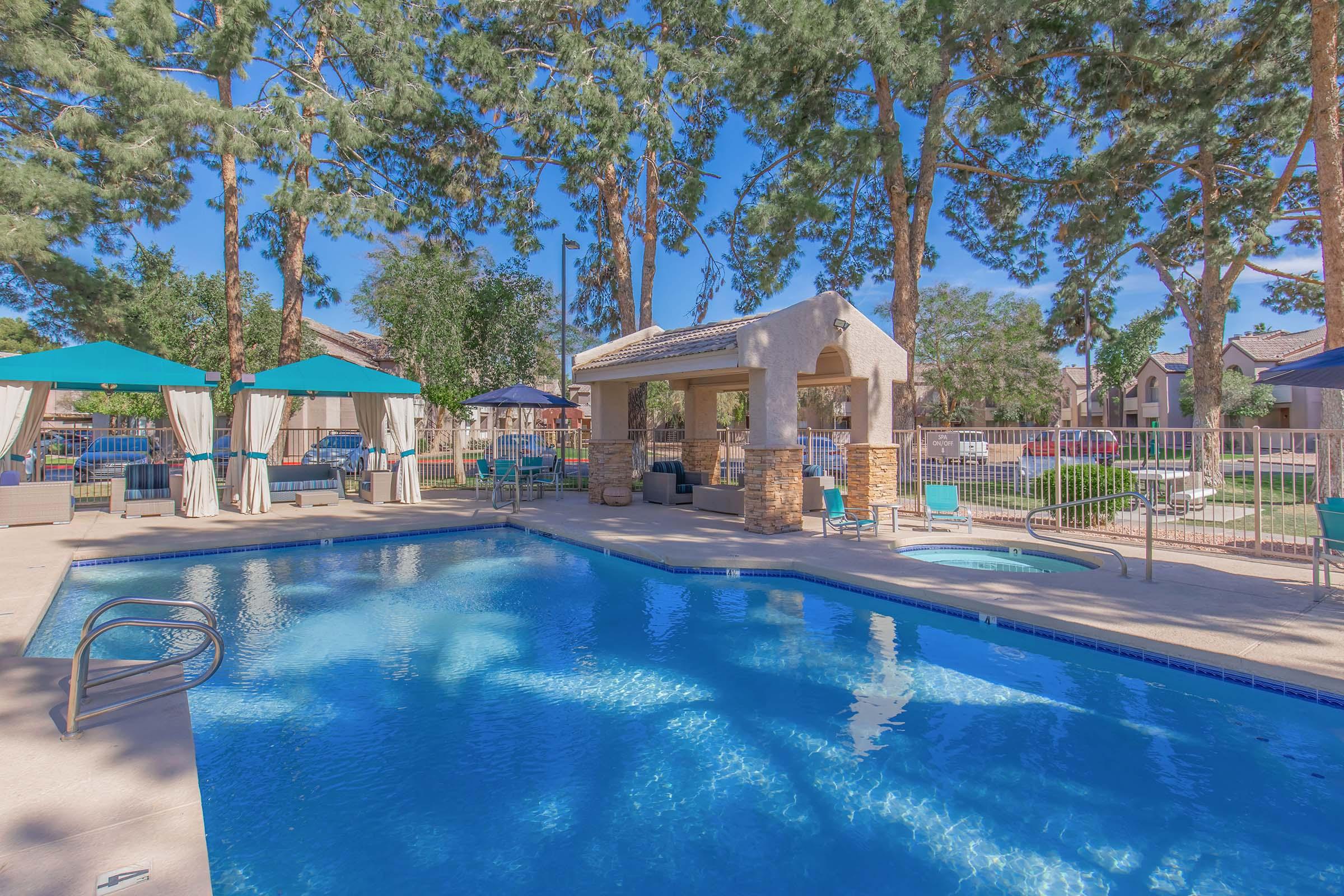 A serene outdoor pool area surrounded by tall trees, featuring shaded lounge spots with umbrellas, comfortable seating, and a small gazebo. The clear blue water reflects the sky and greenery, creating a relaxing atmosphere. Ideal for leisure and enjoyment on a sunny day.