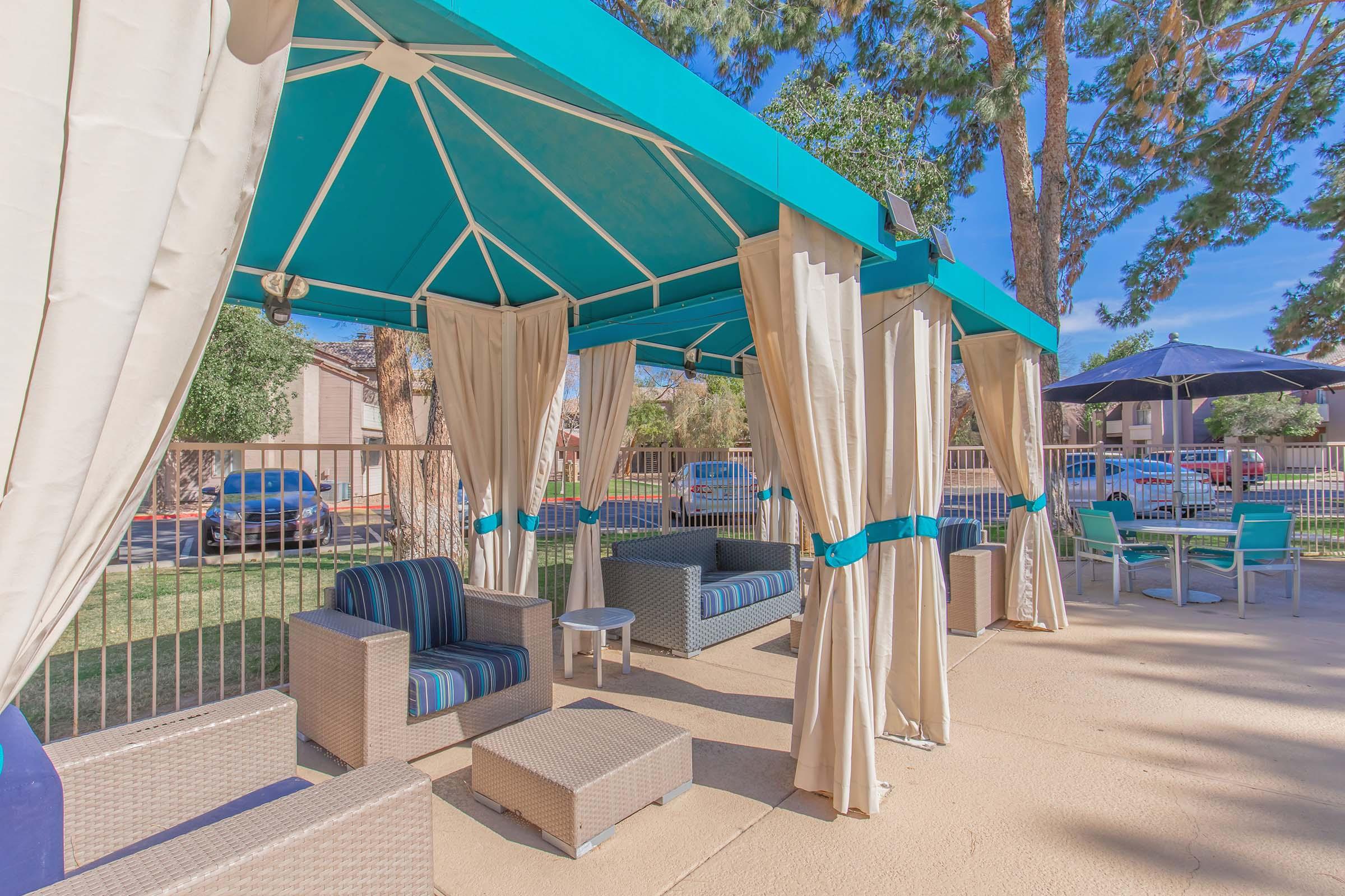 A shaded outdoor seating area featuring a turquoise canopy with curtains, comfortable wicker furniture, and a grass lawn. Nearby, a dining table and umbrellas can be seen, creating a relaxing space for social gatherings.