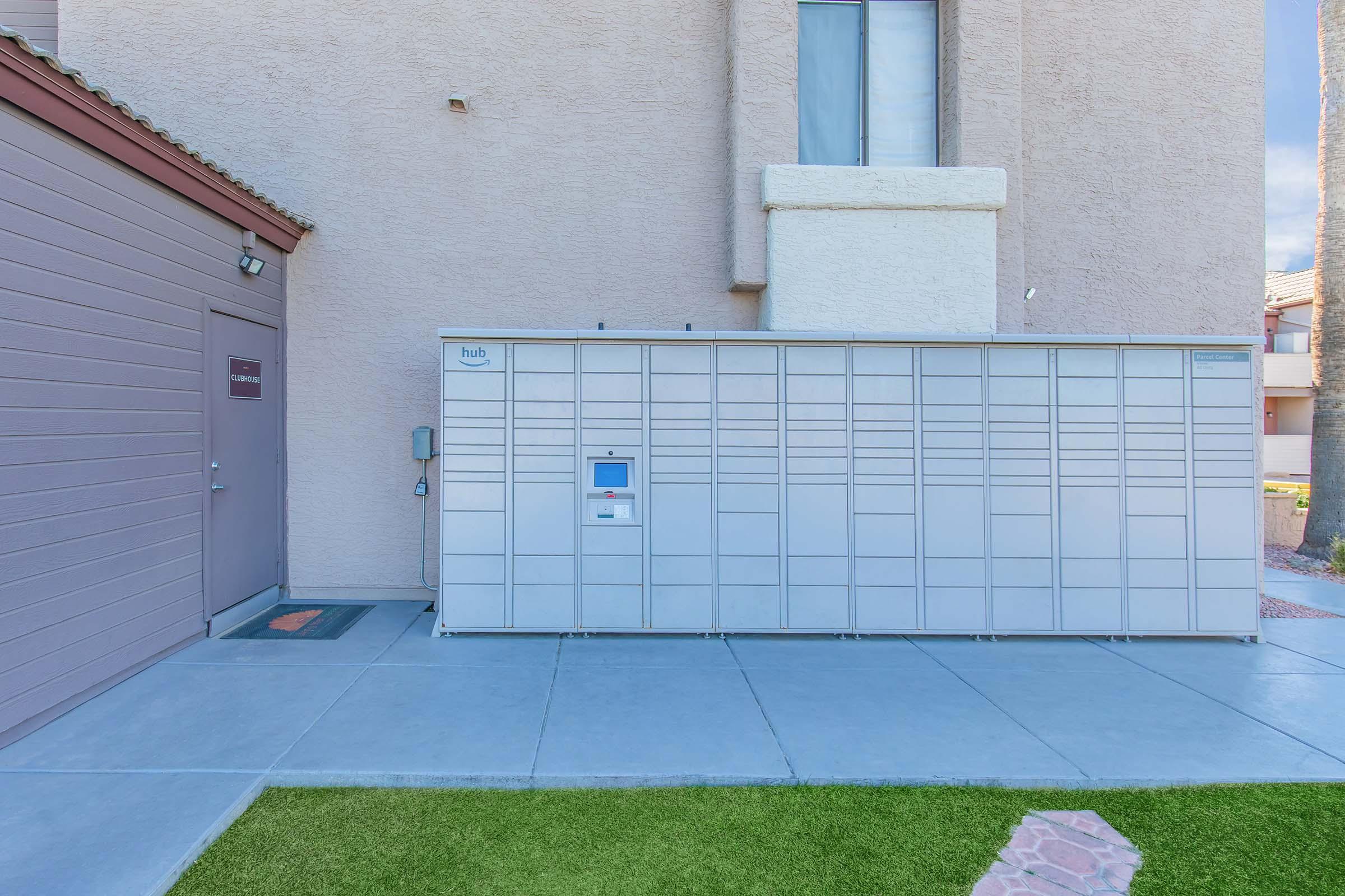 A modern parcel locker station installed outside a building, featuring multiple compartments for package delivery. The structure is metal and gray, with a digital interface for retrieval. Nearby, there's a door to a building with a small welcome mat on the ground and a patch of artificial grass.