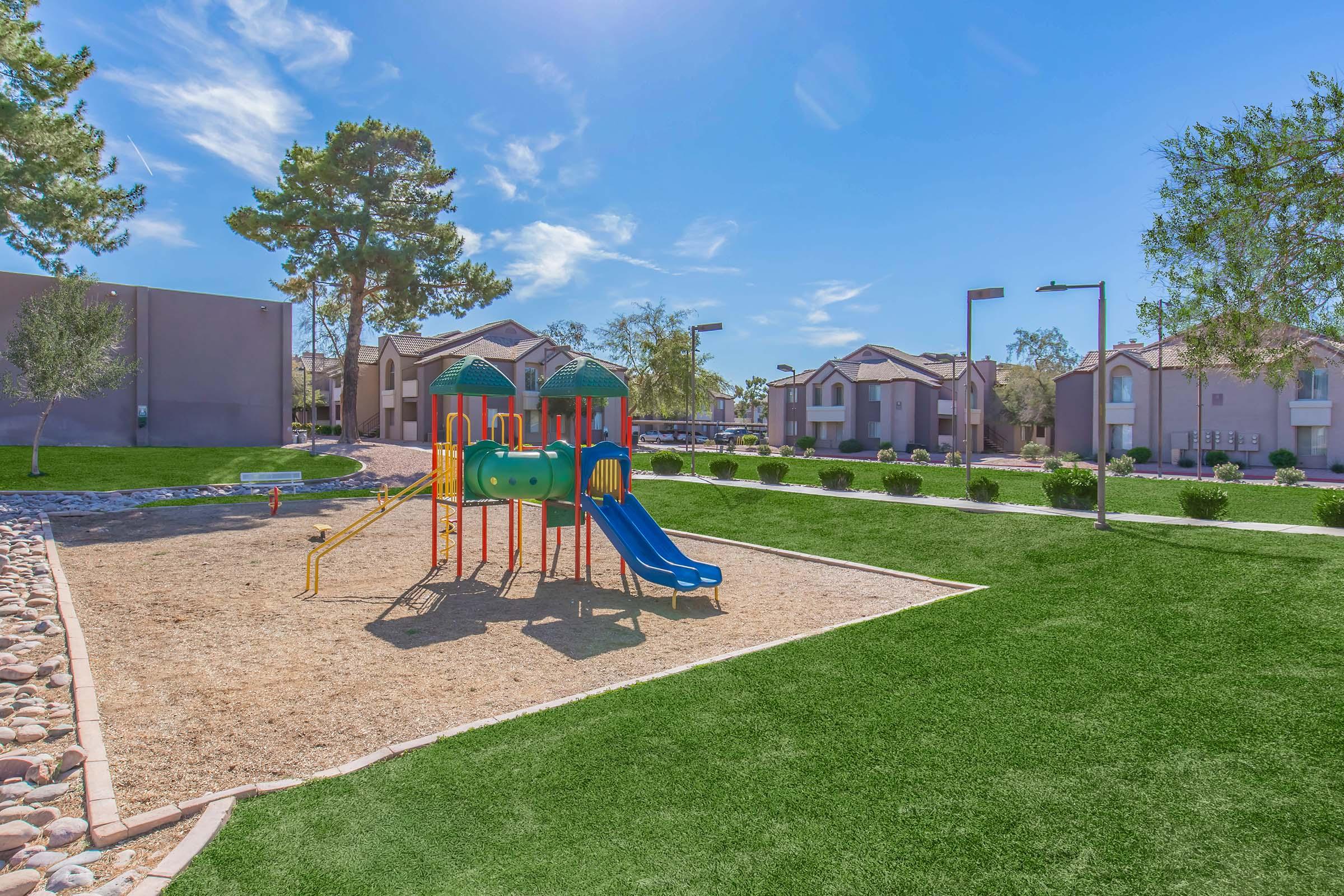 Colorful playground equipment with slides and climbing structures situated on a sandy area, surrounded by green grass and residential buildings. Trees and benches are visible in the background under a clear blue sky.