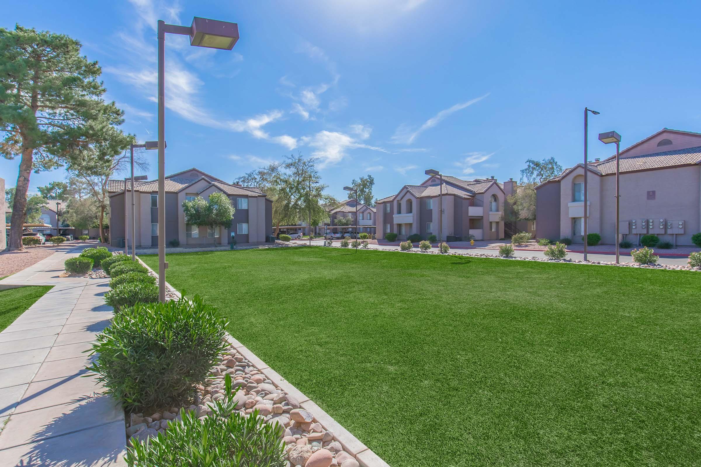 A landscaped courtyard featuring green grass, stone pathways, and decorative shrubs, surrounded by residential buildings. The sky is clear with a few clouds, and there are modern street lamps illuminating the area. The scene conveys a serene and well-maintained outdoor space.