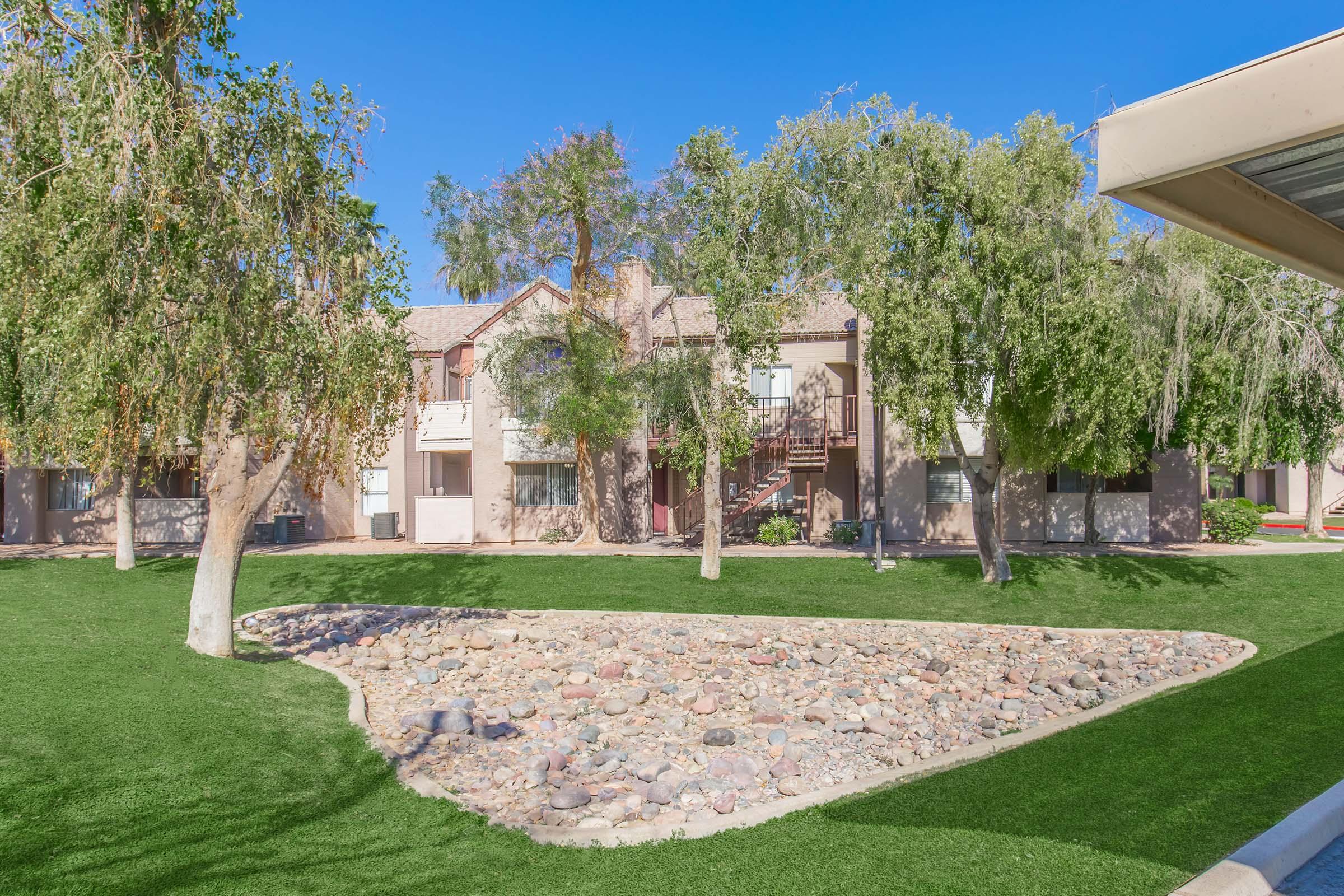 A well-maintained residential complex featuring two-story buildings surrounded by lush green landscaping. In the foreground, a stone-filled area is bordered by manicured grass and trees, creating a serene outdoor environment. Clear blue skies add to the bright and inviting atmosphere.