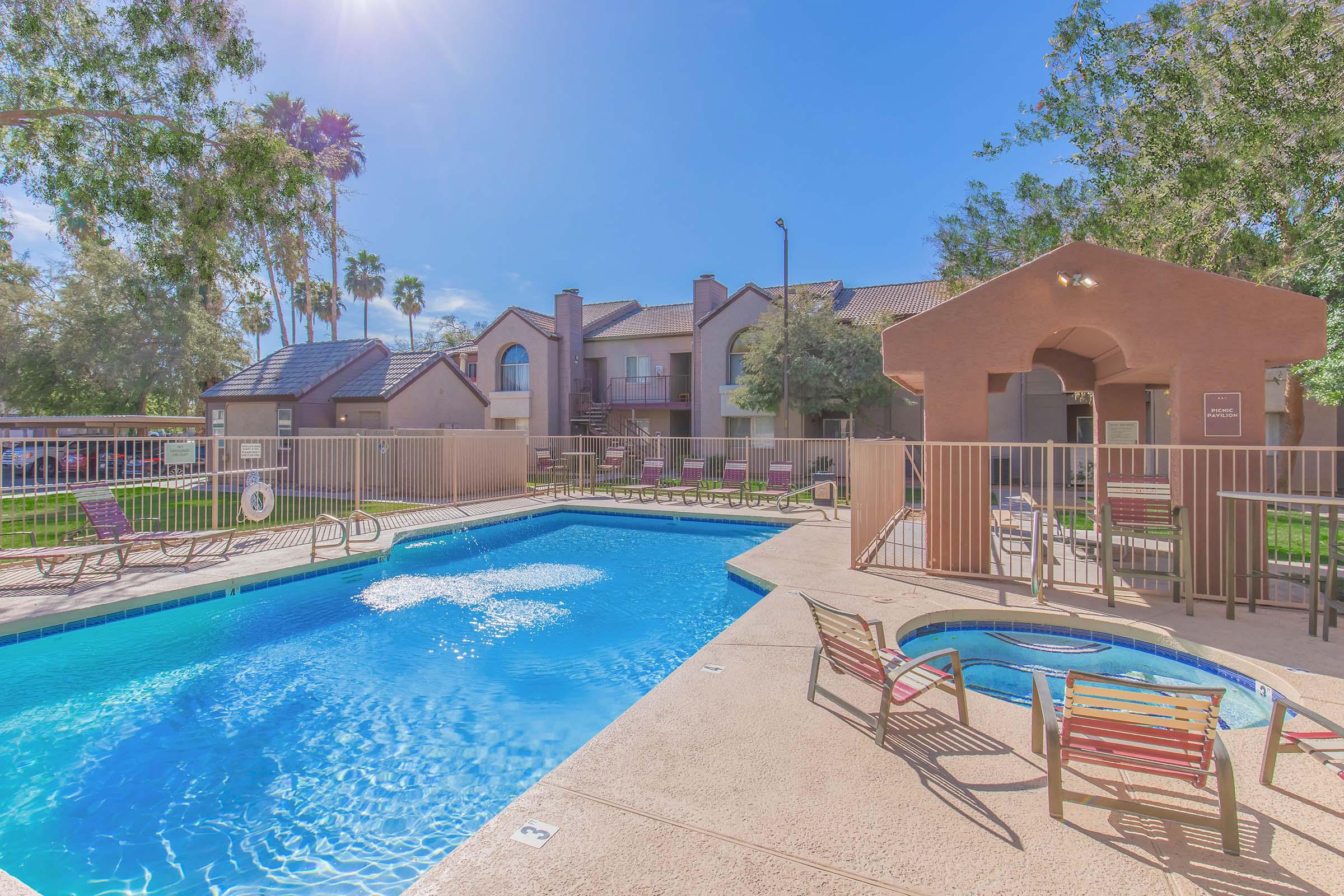 Swimming pool area featuring a clear blue pool and a hot tub. Surrounding the pool are lounge chairs and a shaded seating area. In the background, there are residential buildings and palm trees under a sunny sky.