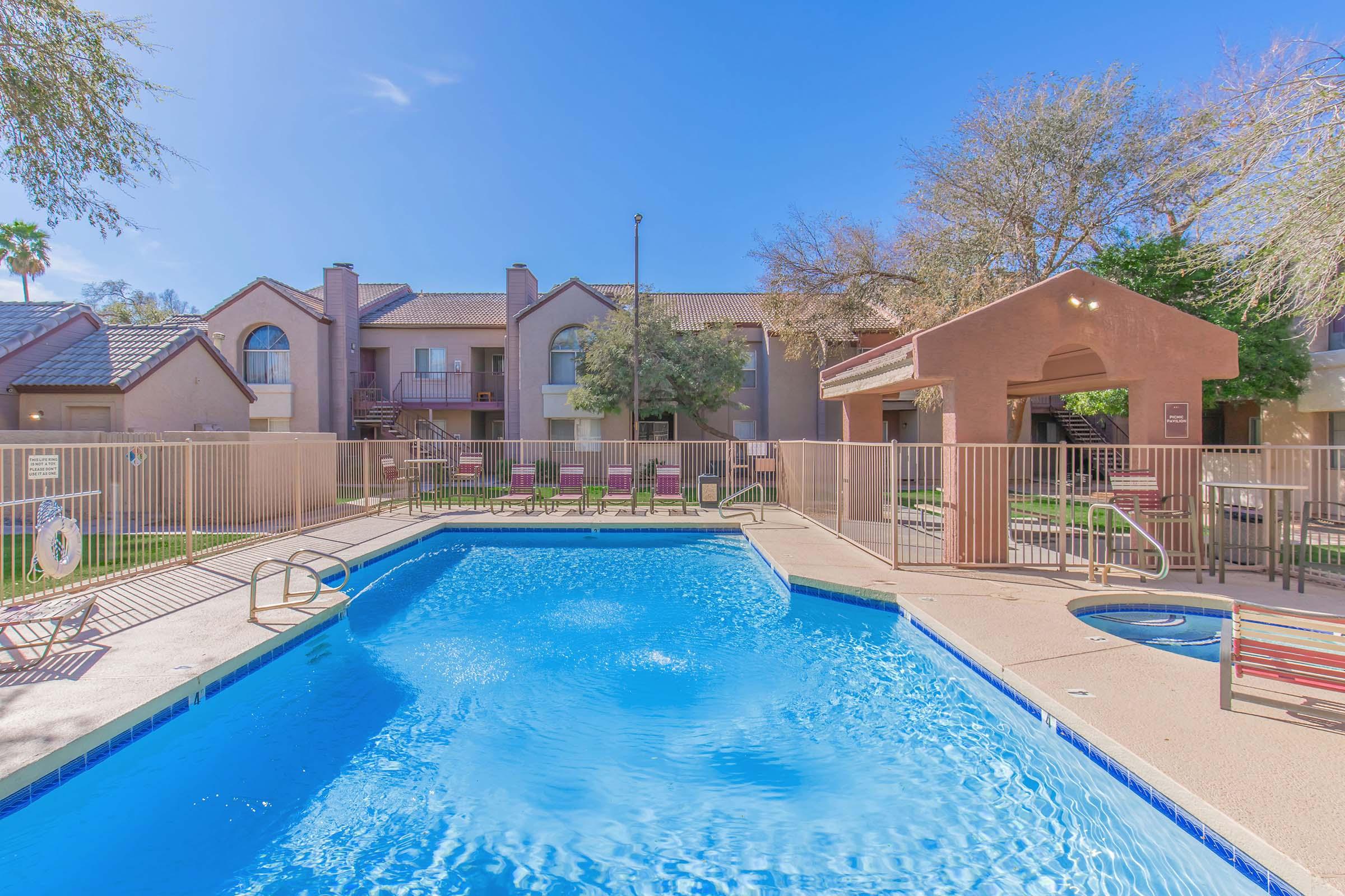 A clear blue swimming pool surrounded by a fenced area, with lounge chairs positioned nearby. In the background, there are two-story apartment buildings with balconies, and trees providing some shade. The sky is bright and sunny, creating a vibrant atmosphere.