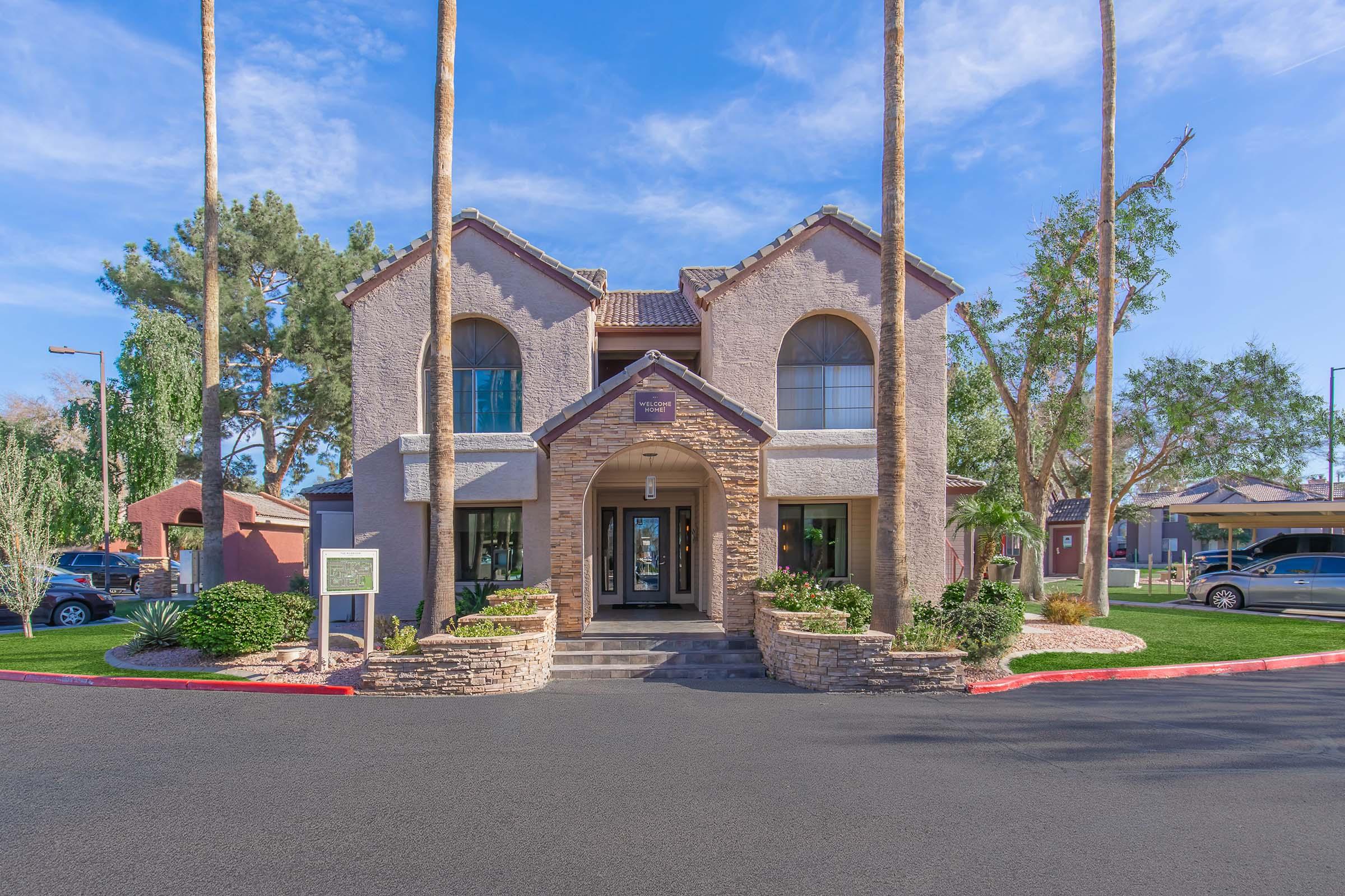 A two-story building with a stone-accented façade, surrounded by palm trees and well-maintained landscaping. The entrance features large windows and double doors, with a light blue sky in the background. A sign is visible in front of the building, and parked cars are seen along the driveway.