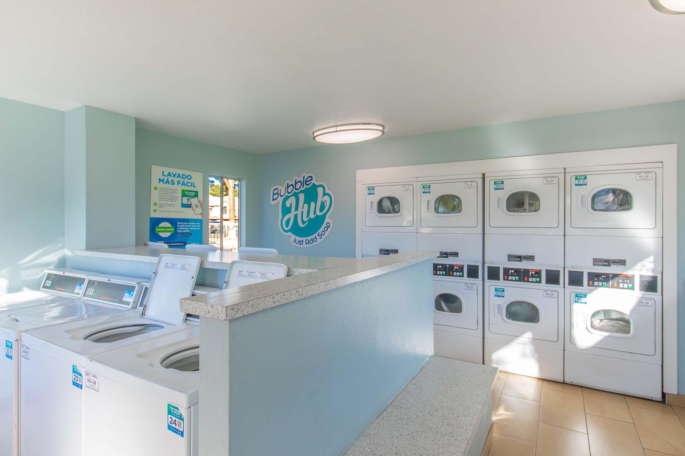 A clean and modern laundry room featuring several washing machines and dryers lined up against a wall. The space has light blue walls, a countertop area, and signage displaying the name "Bubble Hub" and information about laundry services. Natural light brightens the area, creating an inviting atmosphere.