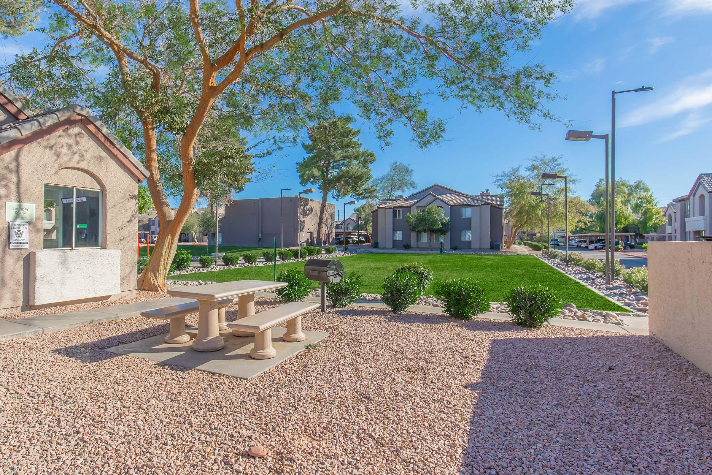 A landscaped area featuring a picnic table surrounded by gravel, shrubs, and trees. In the background, there are residential buildings and a well-maintained lawn. The sky is blue with a few clouds, and there are solar lights positioned along the pathways.