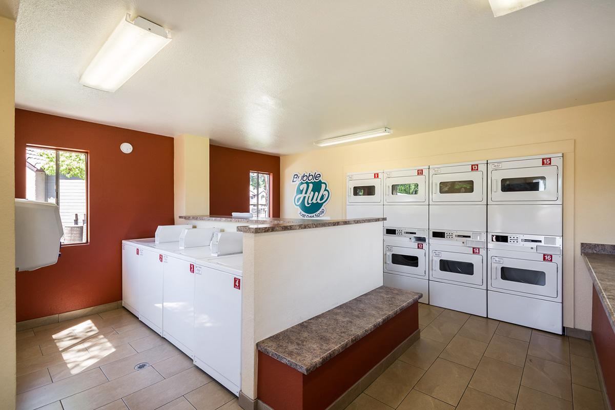 A laundry room featuring several white washing machines and dryers arranged along the wall, with a countertop workspace. The space has bright, overhead lighting, beige walls, and a reddish accent wall. A sign reading "Laundry Hub" is visible in the center. Windows allow natural light in.