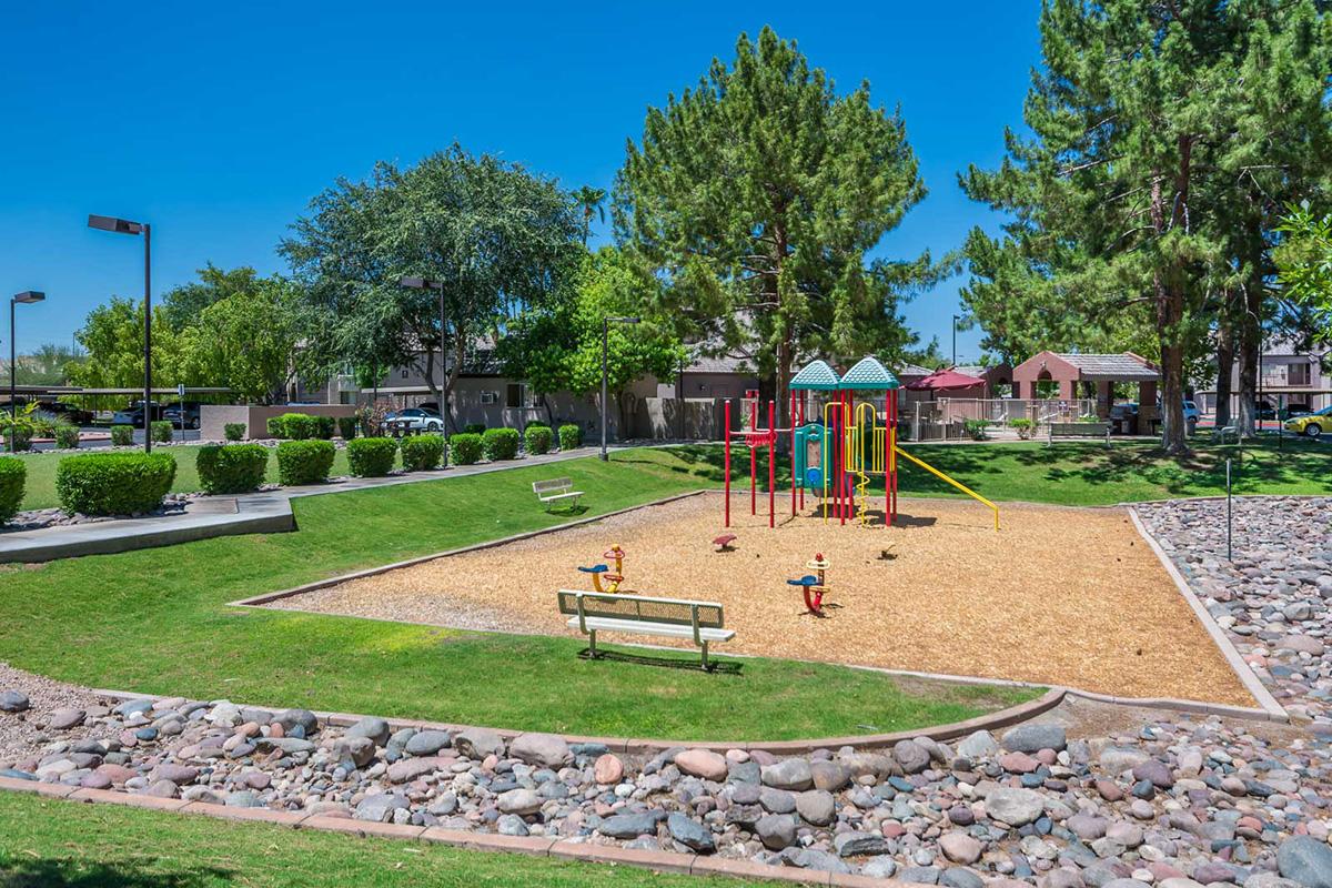 A sunny playground featuring colorful climbing equipment and slides surrounded by green grass and trees. Benches are placed nearby, and the ground is covered with wood chips. The area is well-maintained, with shrubs and a rocky border along the playground.