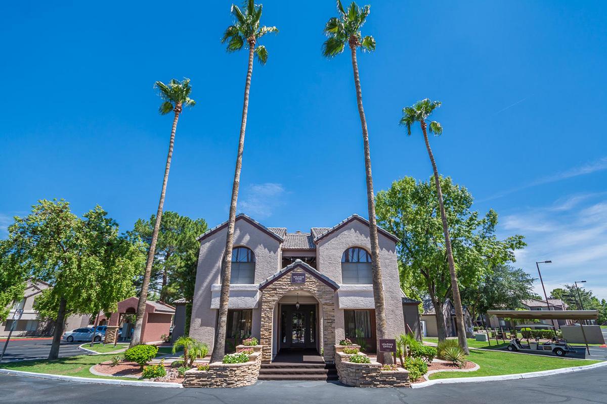 A two-story building with a modern design, featuring large windows and a stone facade, surrounded by tall palm trees and lush greenery. The clear blue sky adds to the bright and welcoming atmosphere, with a landscaped entrance and well-maintained grounds, providing a sense of tranquility and appeal.
