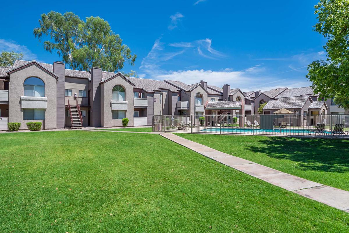 A modern apartment complex with multiple buildings, featuring a well-maintained green lawn and a walkway. There are trees nearby, and a clear blue sky overhead. A swimming pool area is visible behind the buildings, surrounded by a fence.