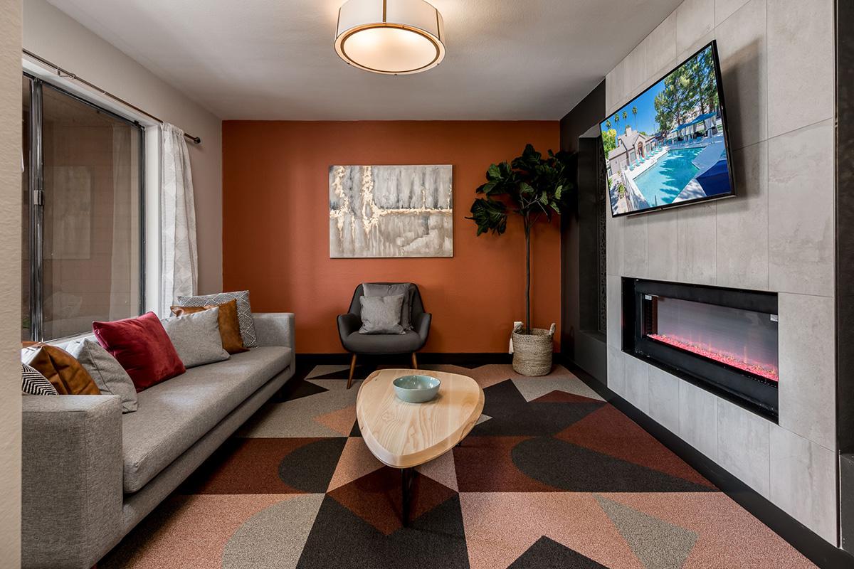 A modern living room featuring a gray sofa with colorful throw pillows, a wooden coffee table, and a wall-mounted TV displaying a pool scene. The room has an orange accent wall, a potted plant, artwork, and a contemporary electric fireplace, enhancing the cozy atmosphere.